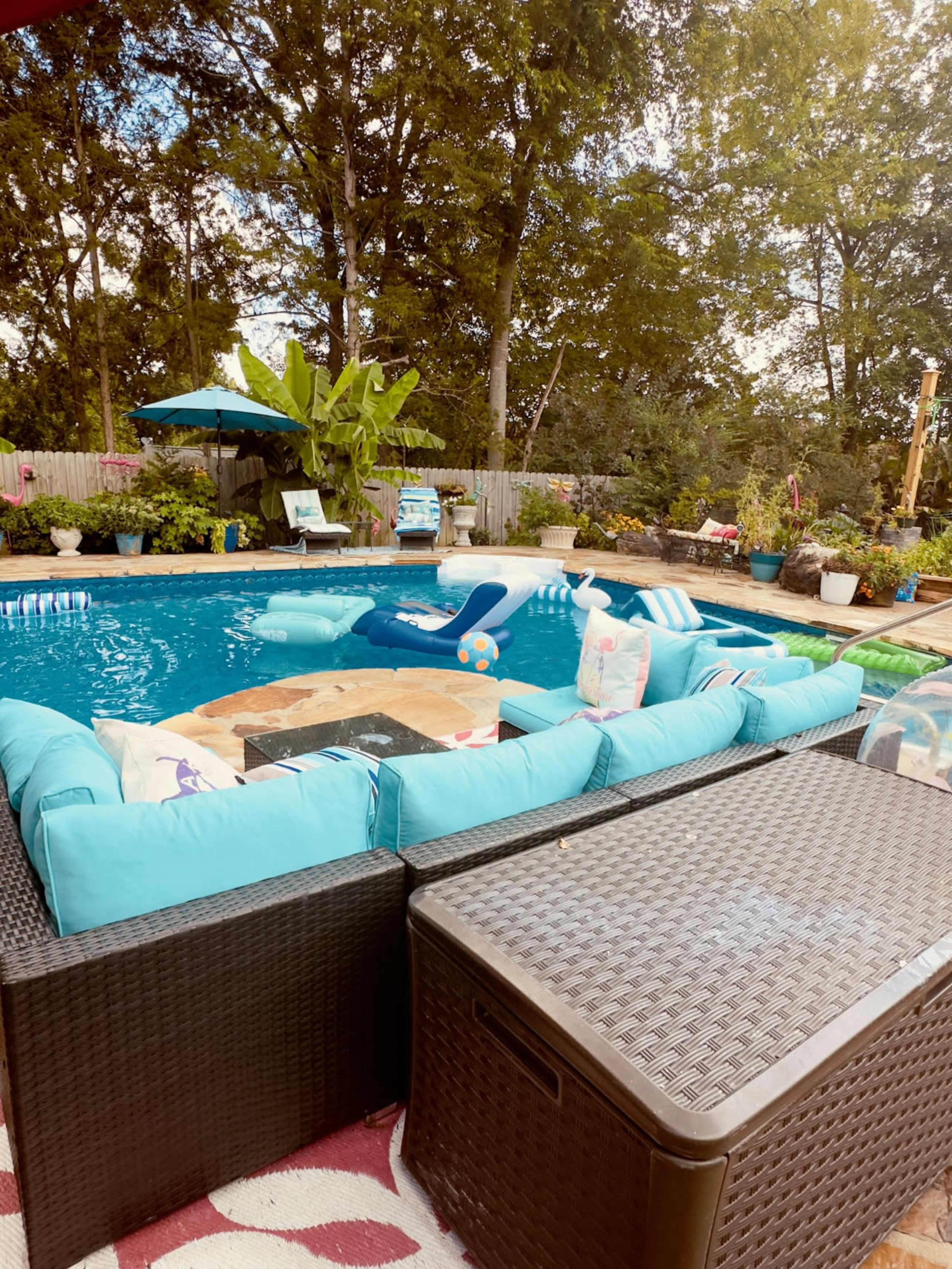 A brightly colored pool area features a lounge with turquoise cushions and a table, surrounded by greenery and decorative plants.