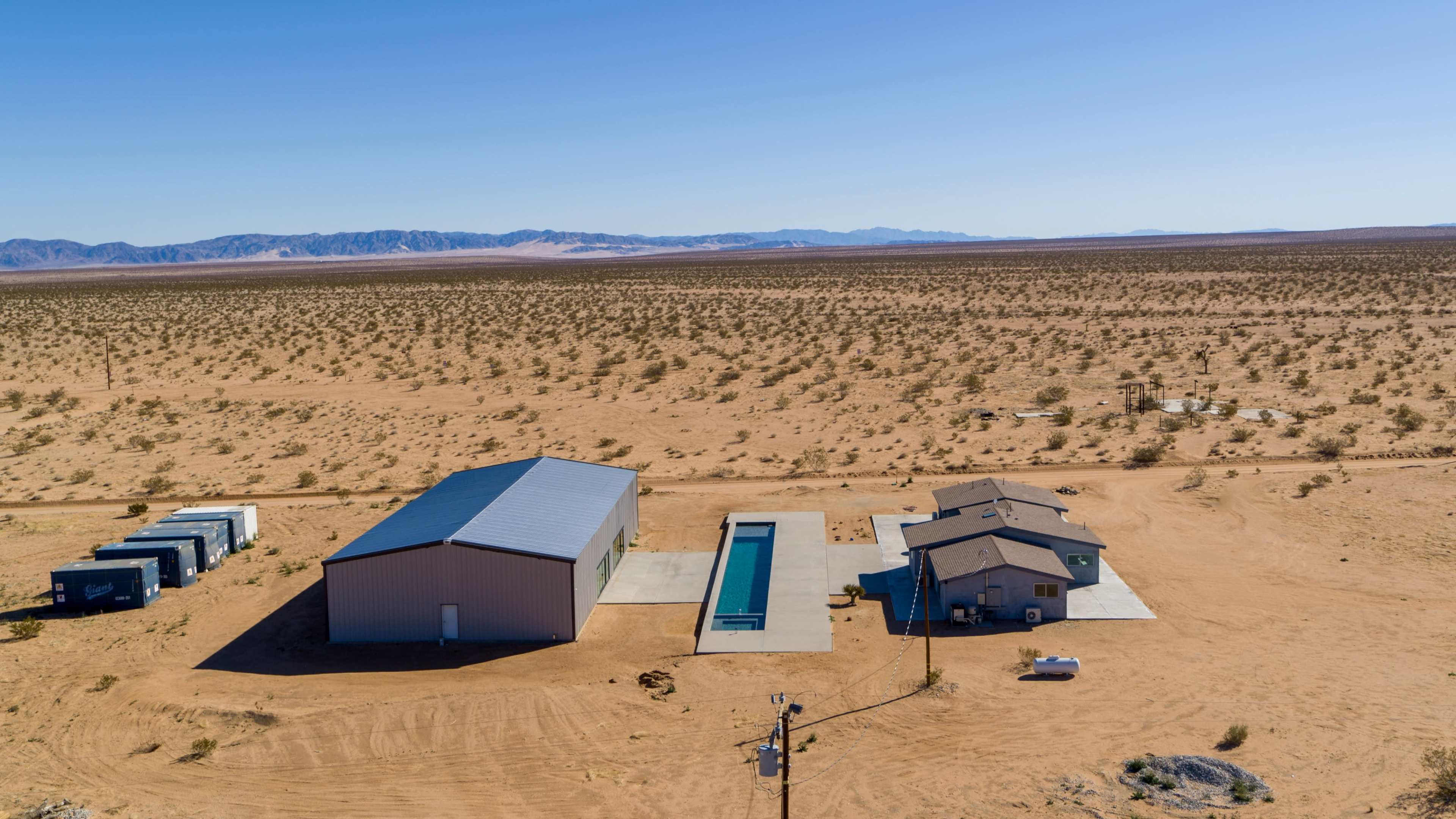 A desert landscape with a large metal building, a swimming pool, and a house situated on sandy terrain, surrounded by sparse vegetation and distant mountains.