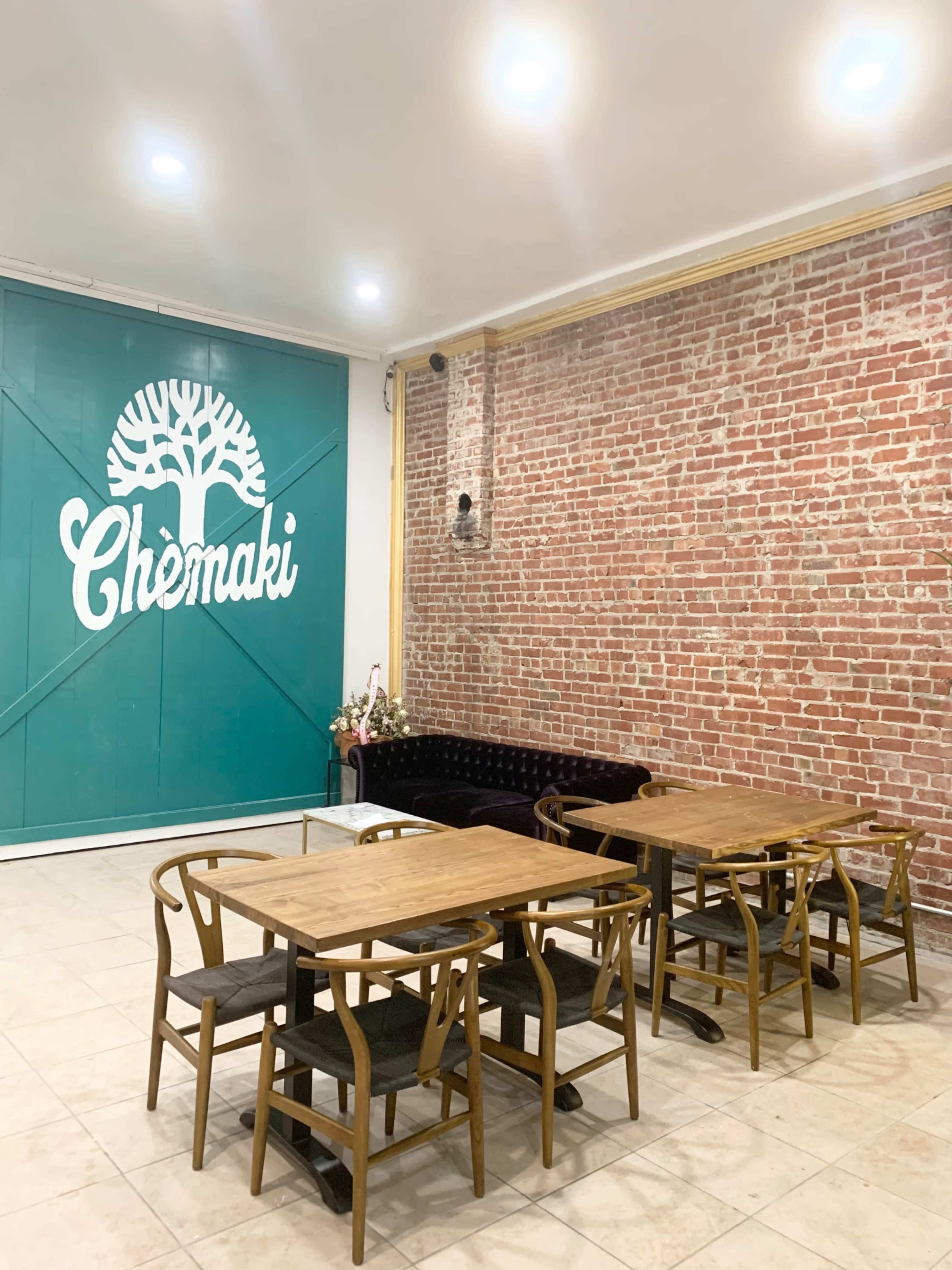 A cozy café interior features wooden tables and chairs set against a brick wall, with a large turquoise logo on one side.