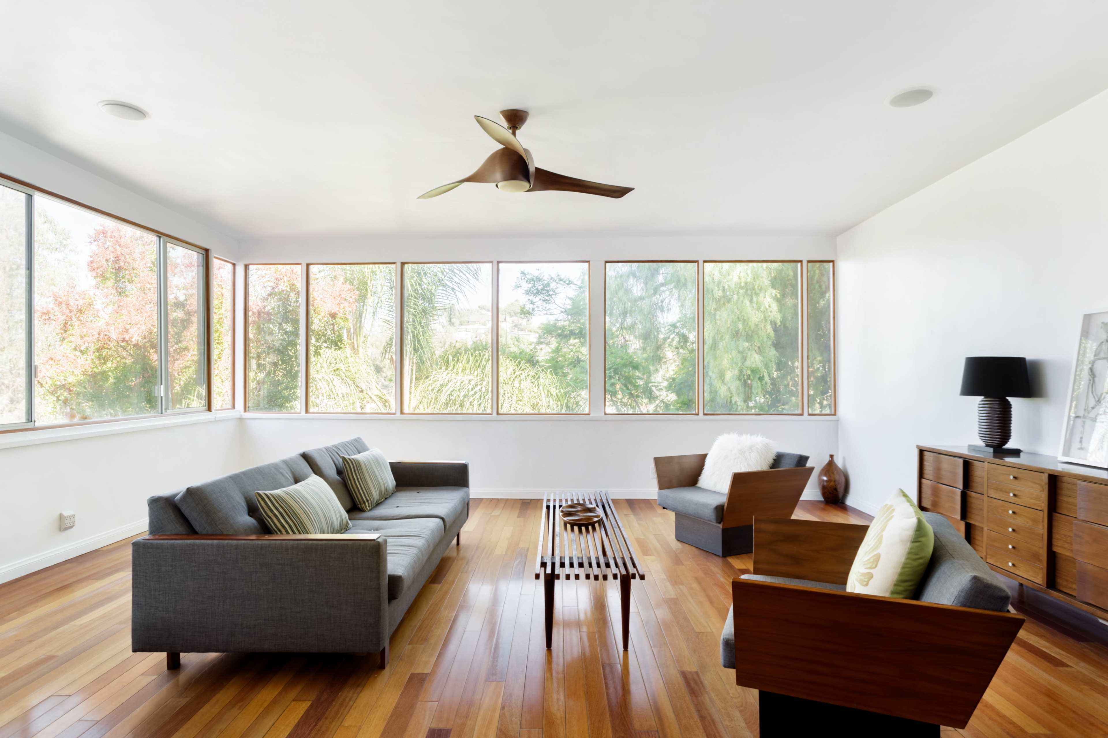 A modern living room features a gray sofa and armchairs around a wooden coffee table, with large windows allowing natural light and views of greenery.