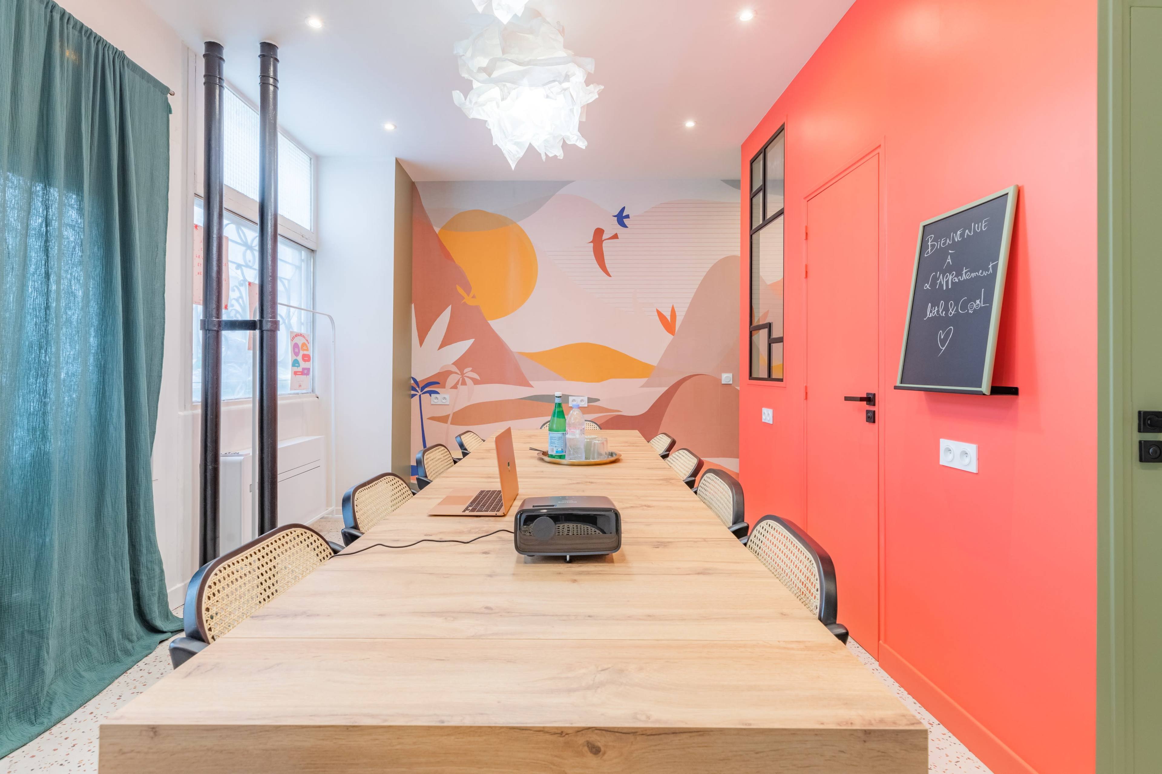 A long wooden table with a laptop and a toaster sits in a brightly colored room featuring a mural on one wall and a chalkboard on another.