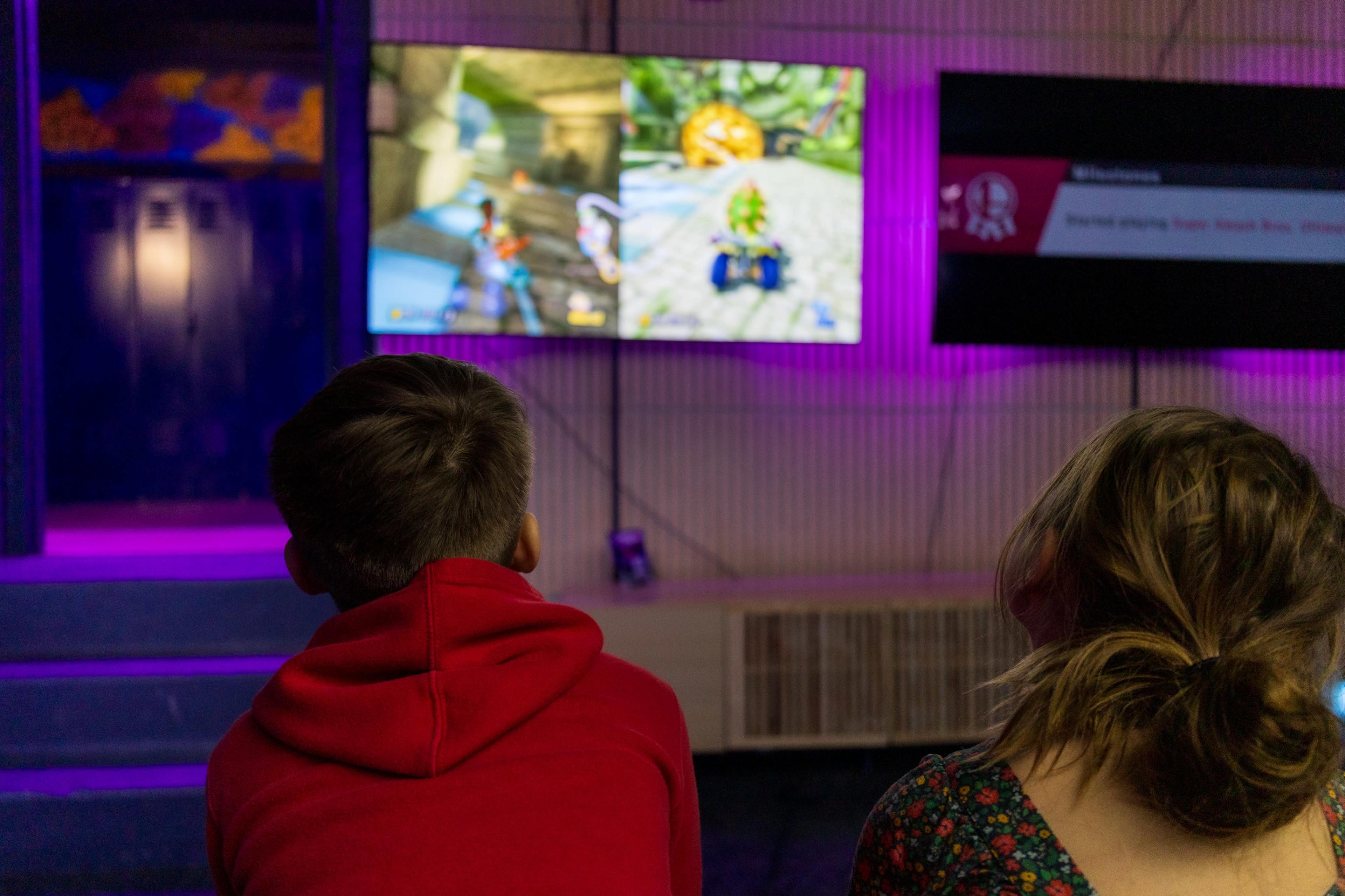 Two children watch a video game being played on a large screen in a dimly lit room.