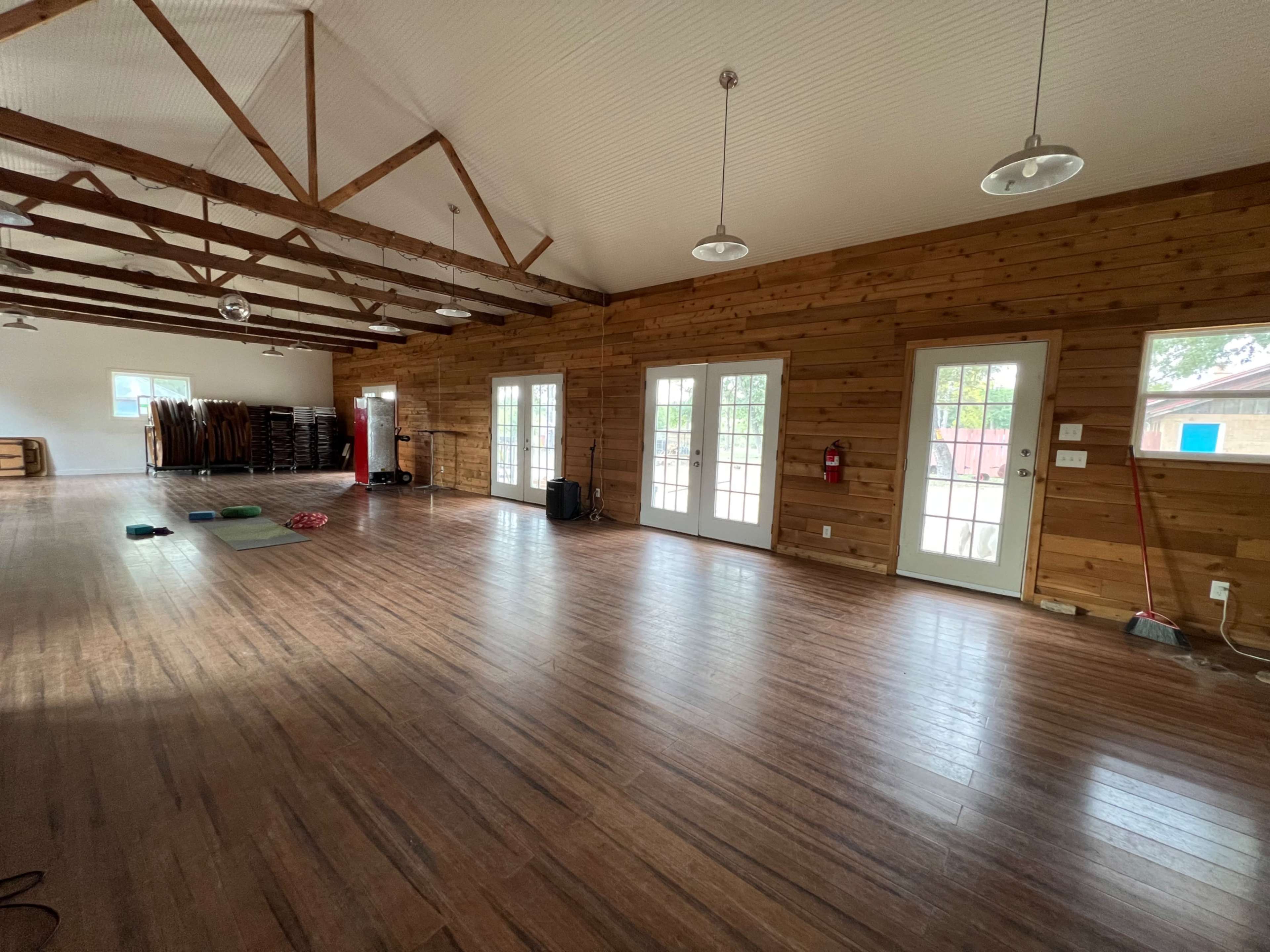 The image shows a spacious room with wooden walls, large windows, and a polished wooden floor, featuring various fitness mats and equipment arranged along one side.