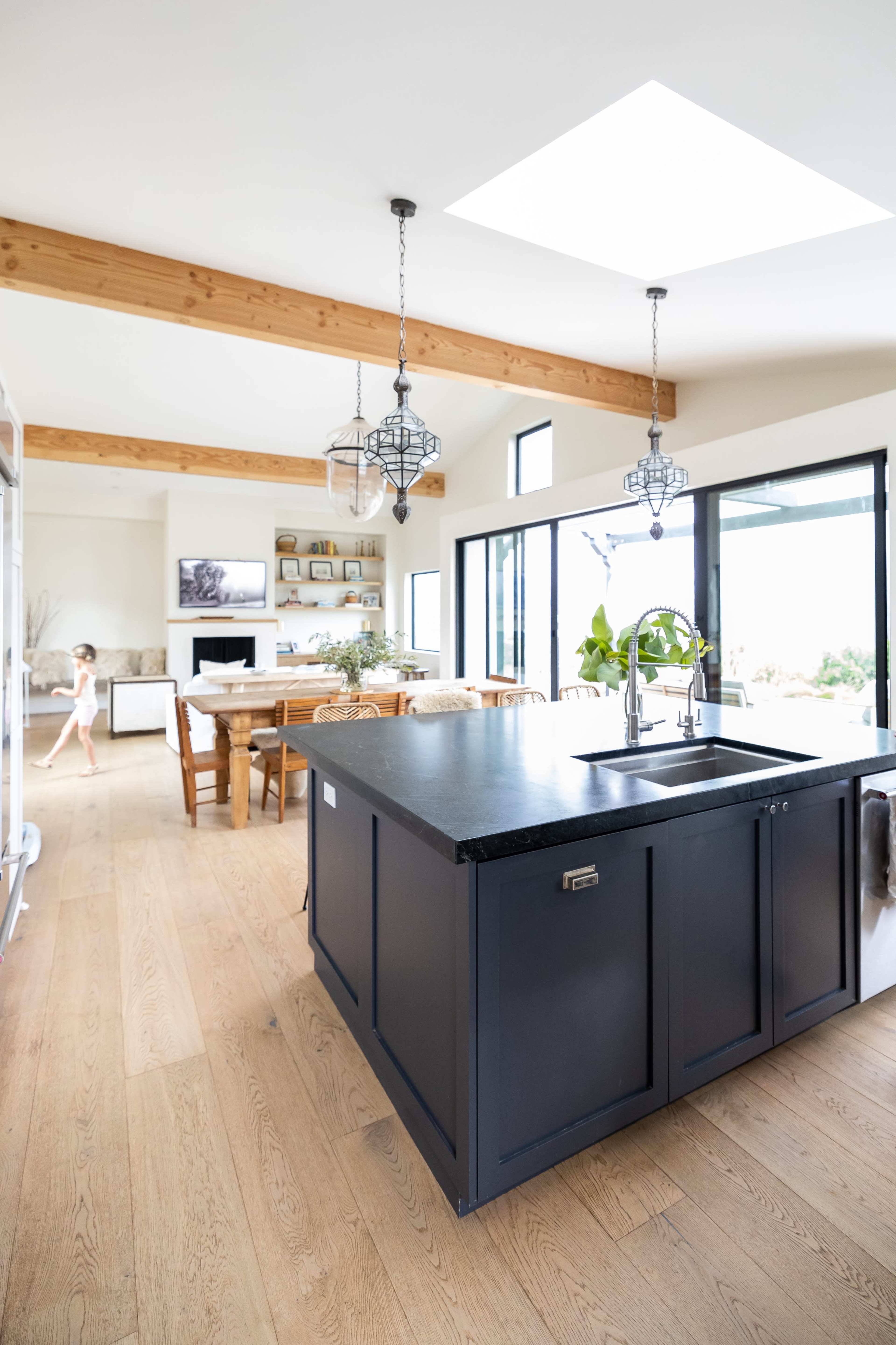 The image shows a modern kitchen featuring a large dark island, a dining area with a wooden table, and large windows that provide ample natural light.