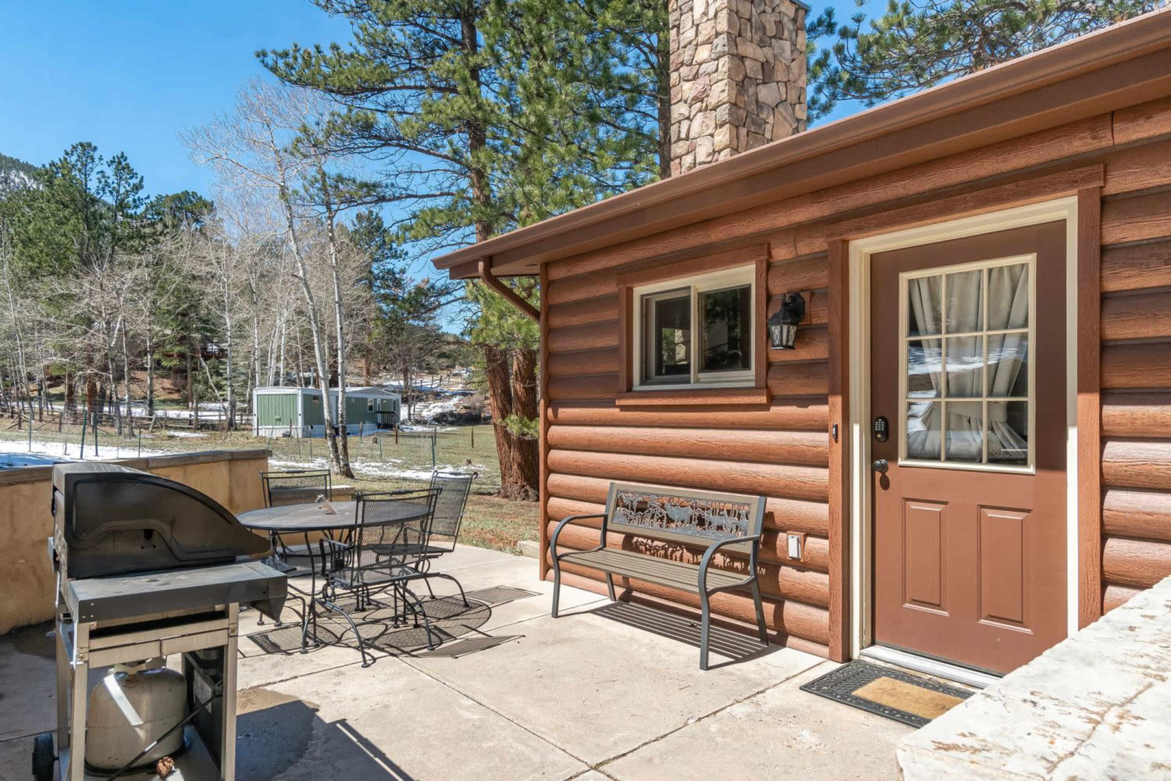 A log cabin features a patio with a grill and a table set for outdoor dining, surrounded by pine trees and patches of snow.