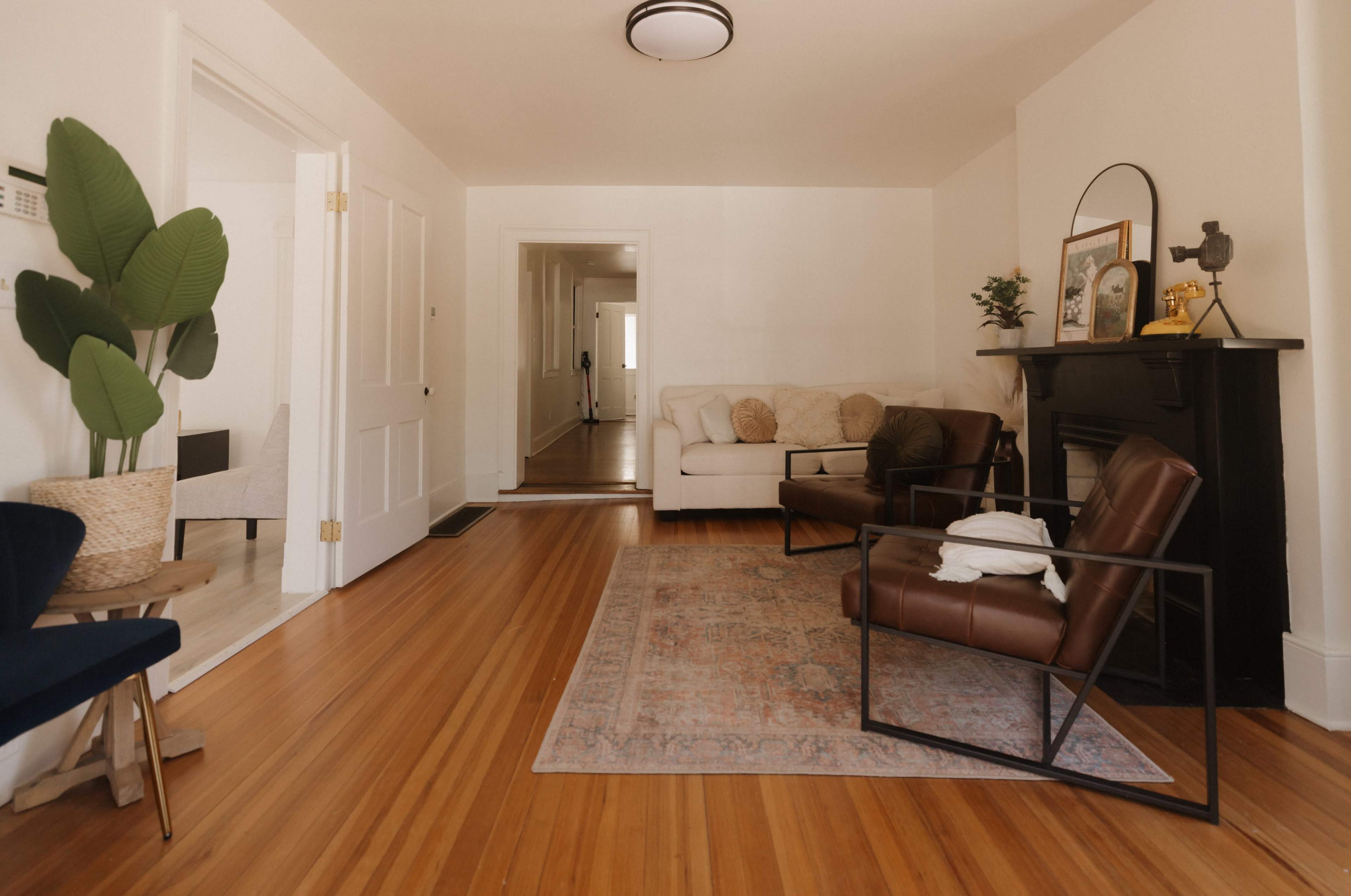 The image shows a living room with hardwood floors, a sofa, two armchairs, and a decorative rug, along with a doorway leading to a hallway.