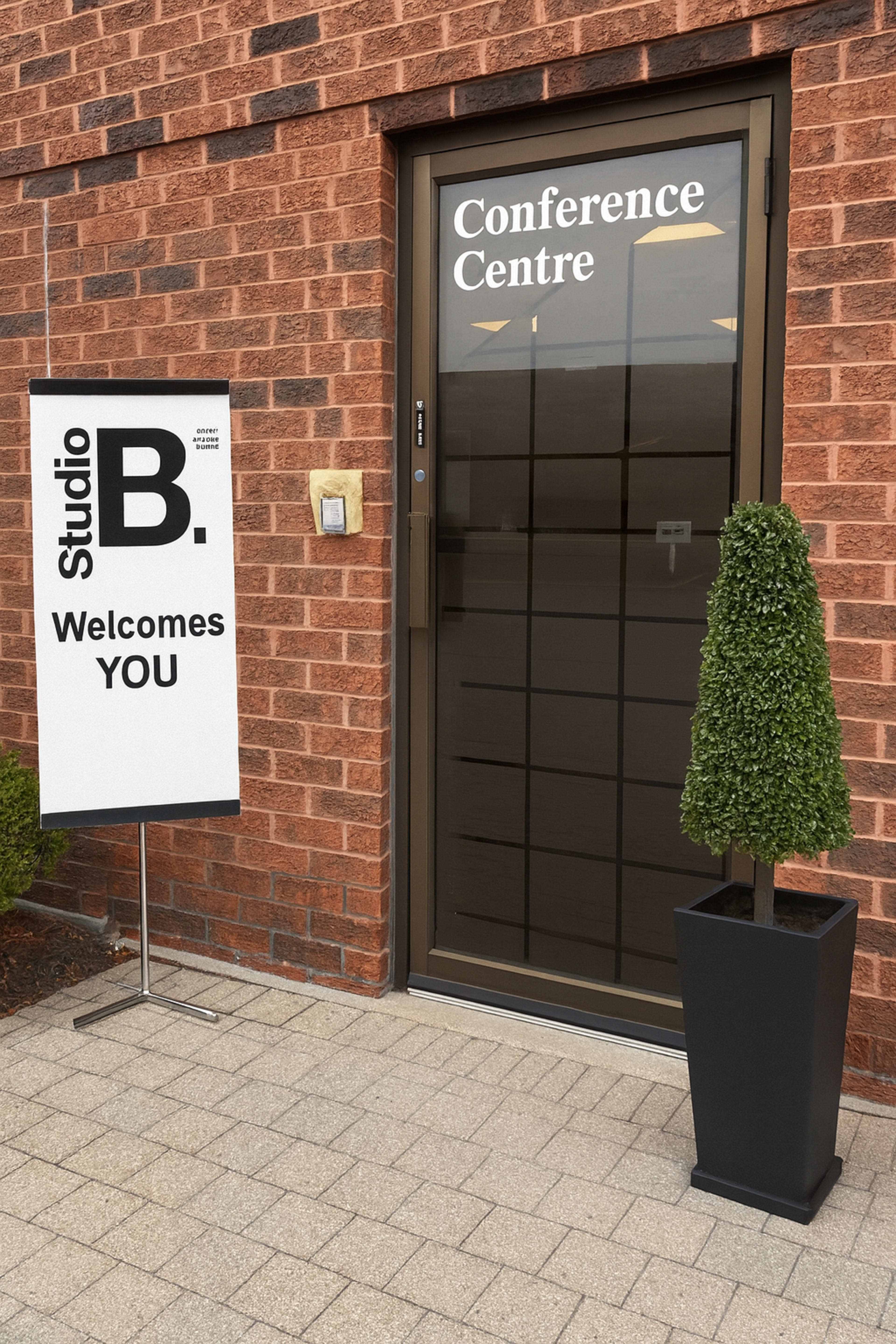 The image shows a conference center entrance with a modern sign displaying "Studio B. Welcomes YOU" next to a neatly trimmed topiary in a planter.