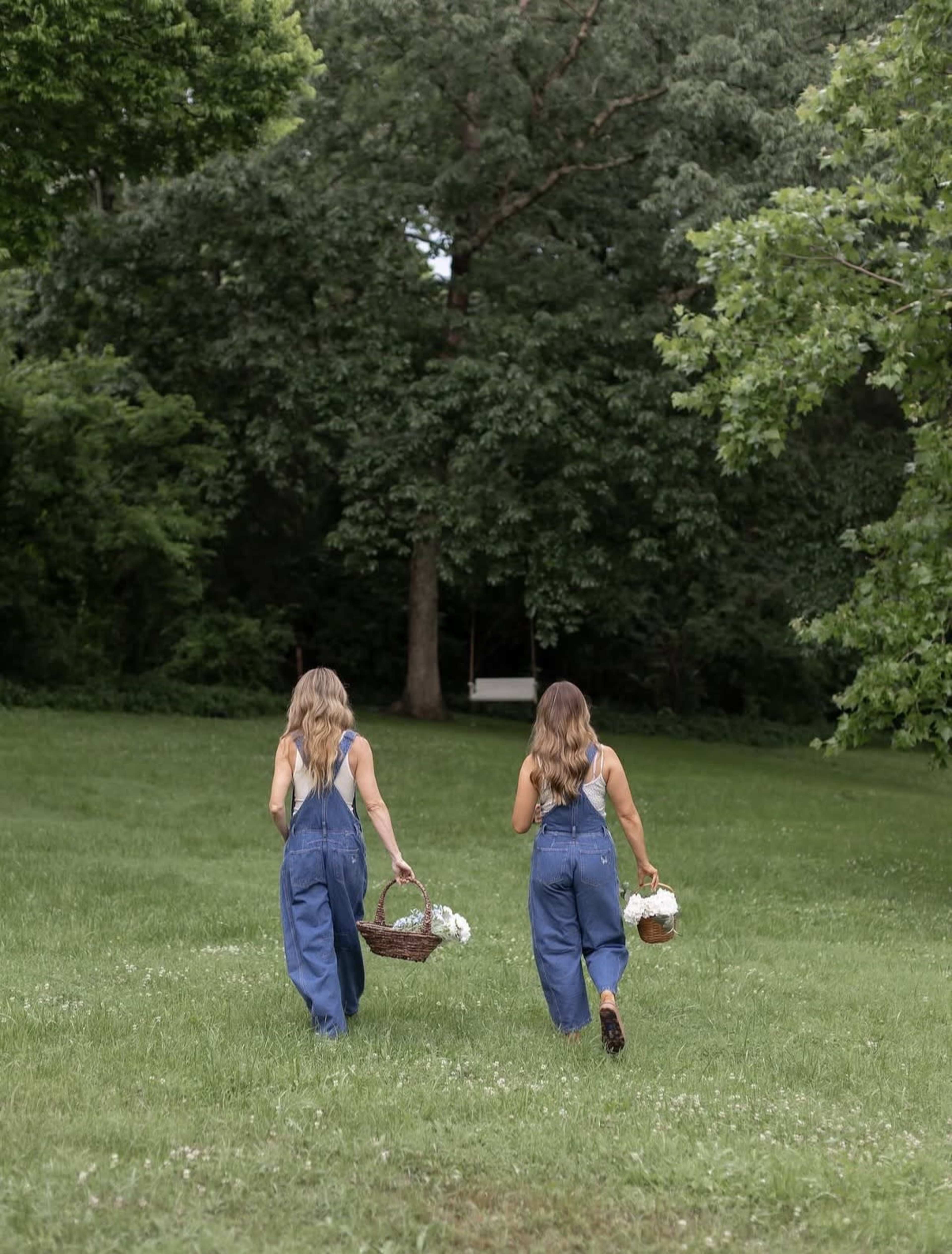 Two women wearing denim overalls walk through a grassy field while carrying wicker baskets.
