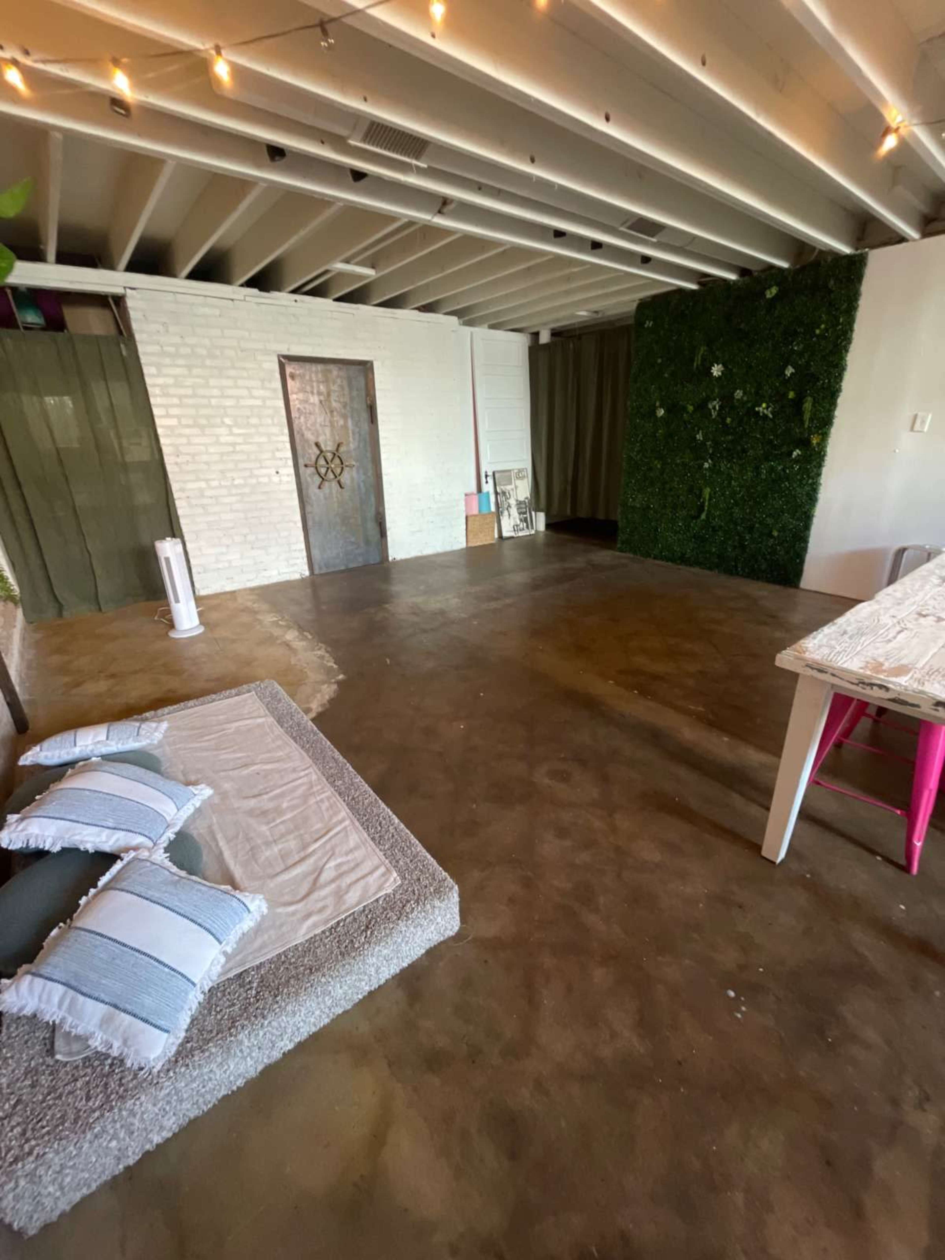 The image shows a spacious, modern interior with a concrete floor, a sectional seating area with pillows, a small table, and a green wall of plants.