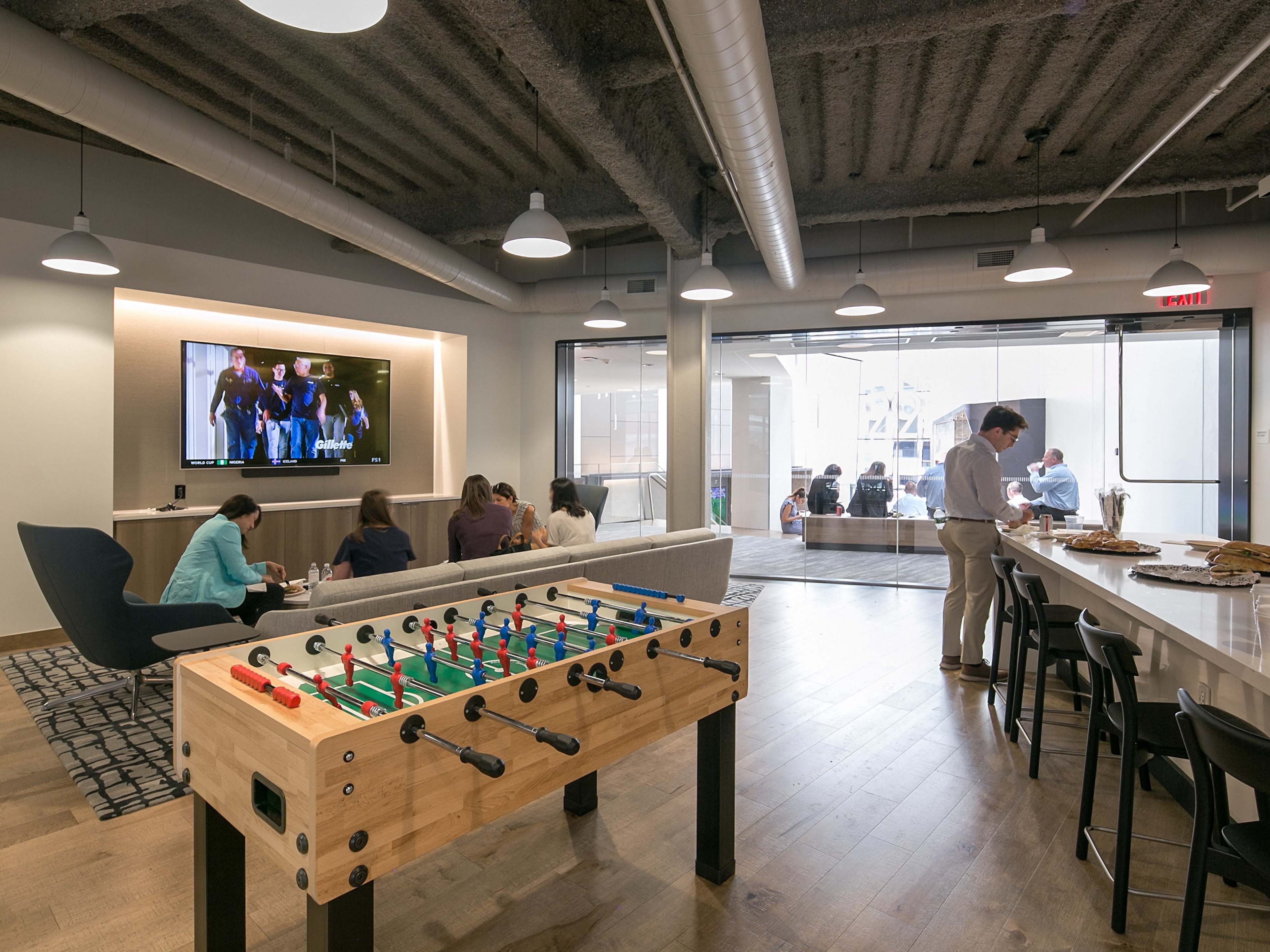 A common area features a foosball table in the foreground, with people engaged in various activities, such as watching a screen and socializing around tables.
