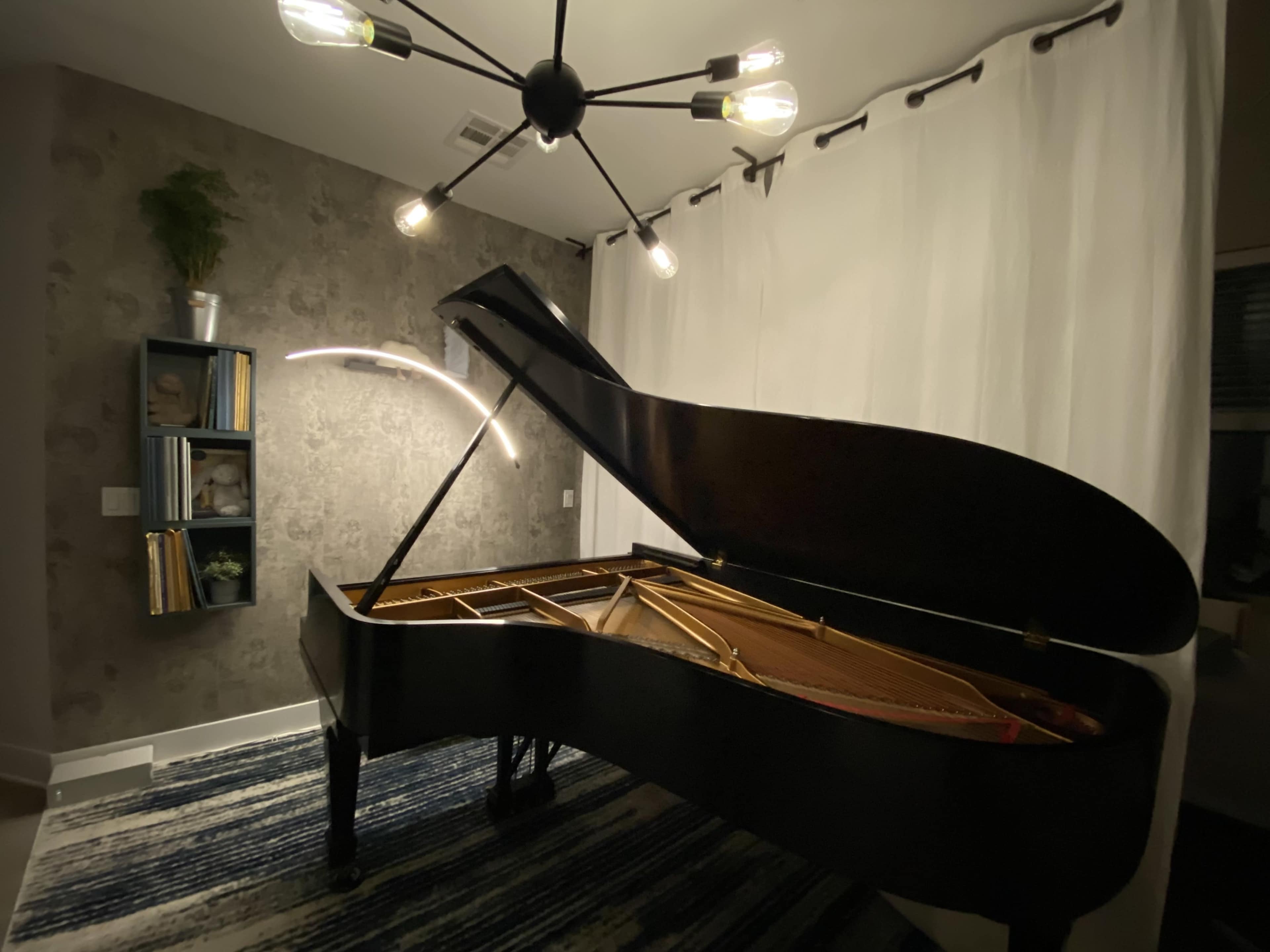 A black grand piano sits in a well-lit room with a textured wall and a modern chandelier above.