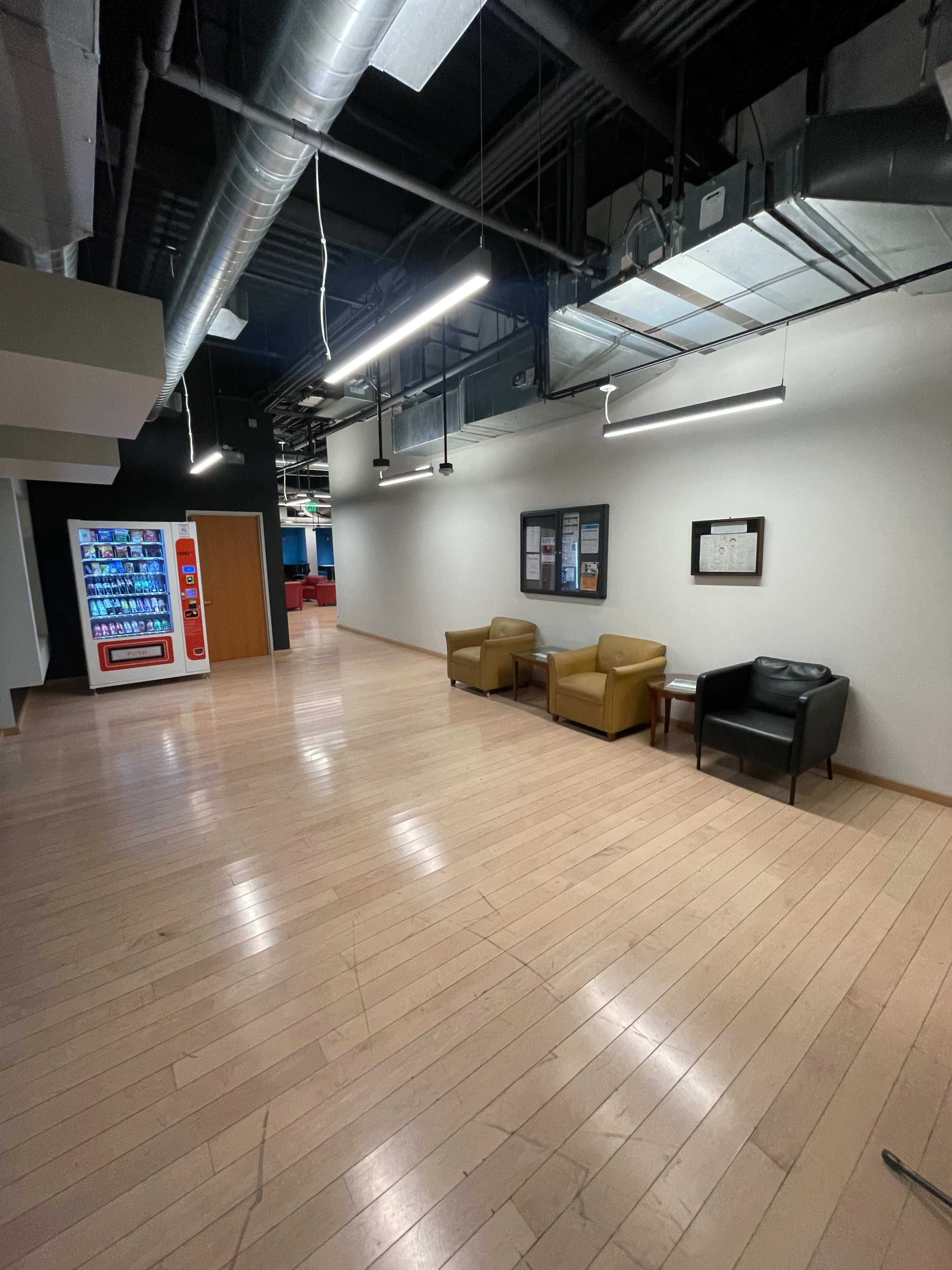 The image shows a spacious hallway with wooden flooring, two chairs in the center, and a vending machine against one wall.