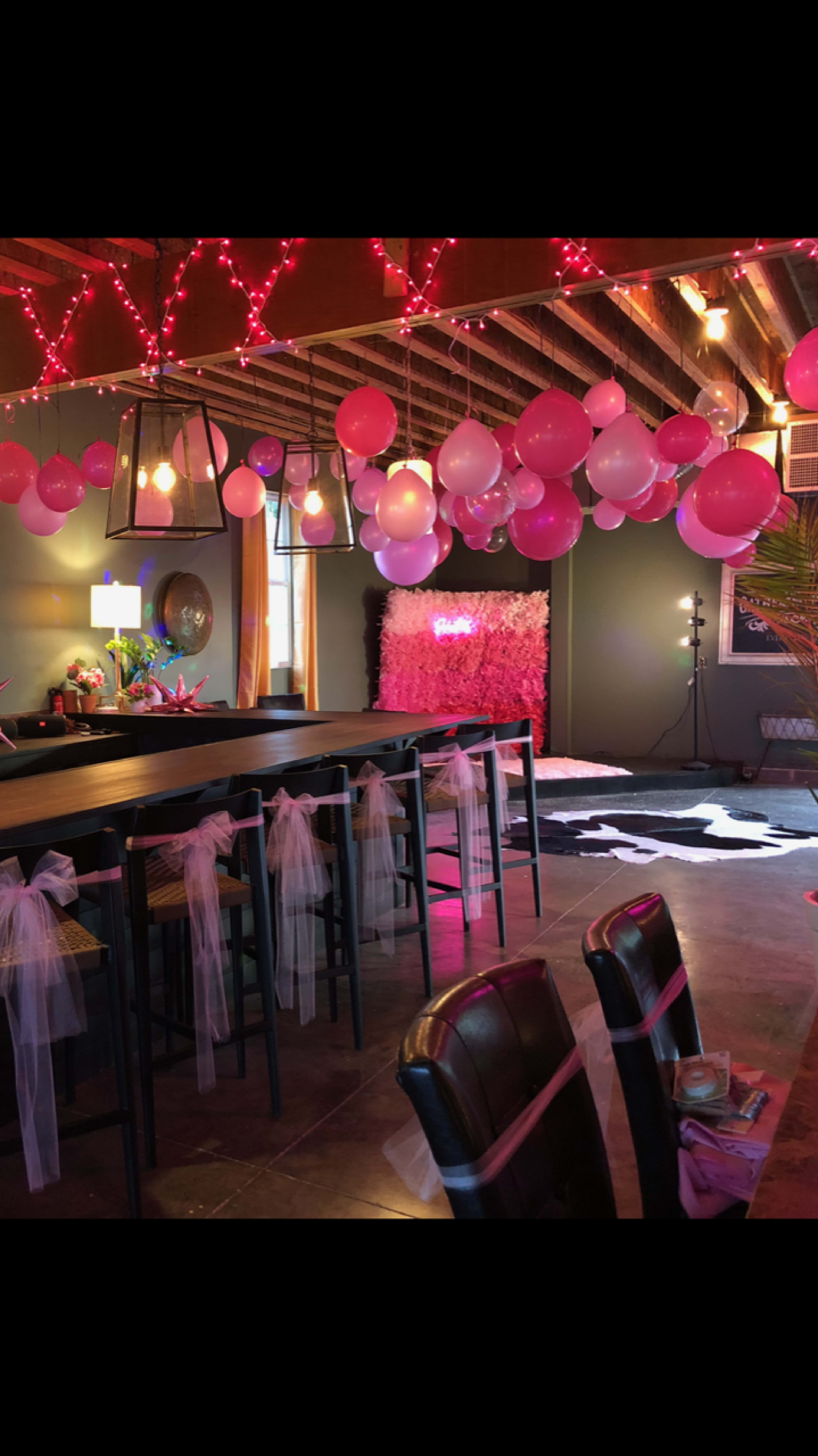 A decorated event space featuring pink balloons and fabric drapes hanging from the ceiling, with a long table adorned with sashes.
