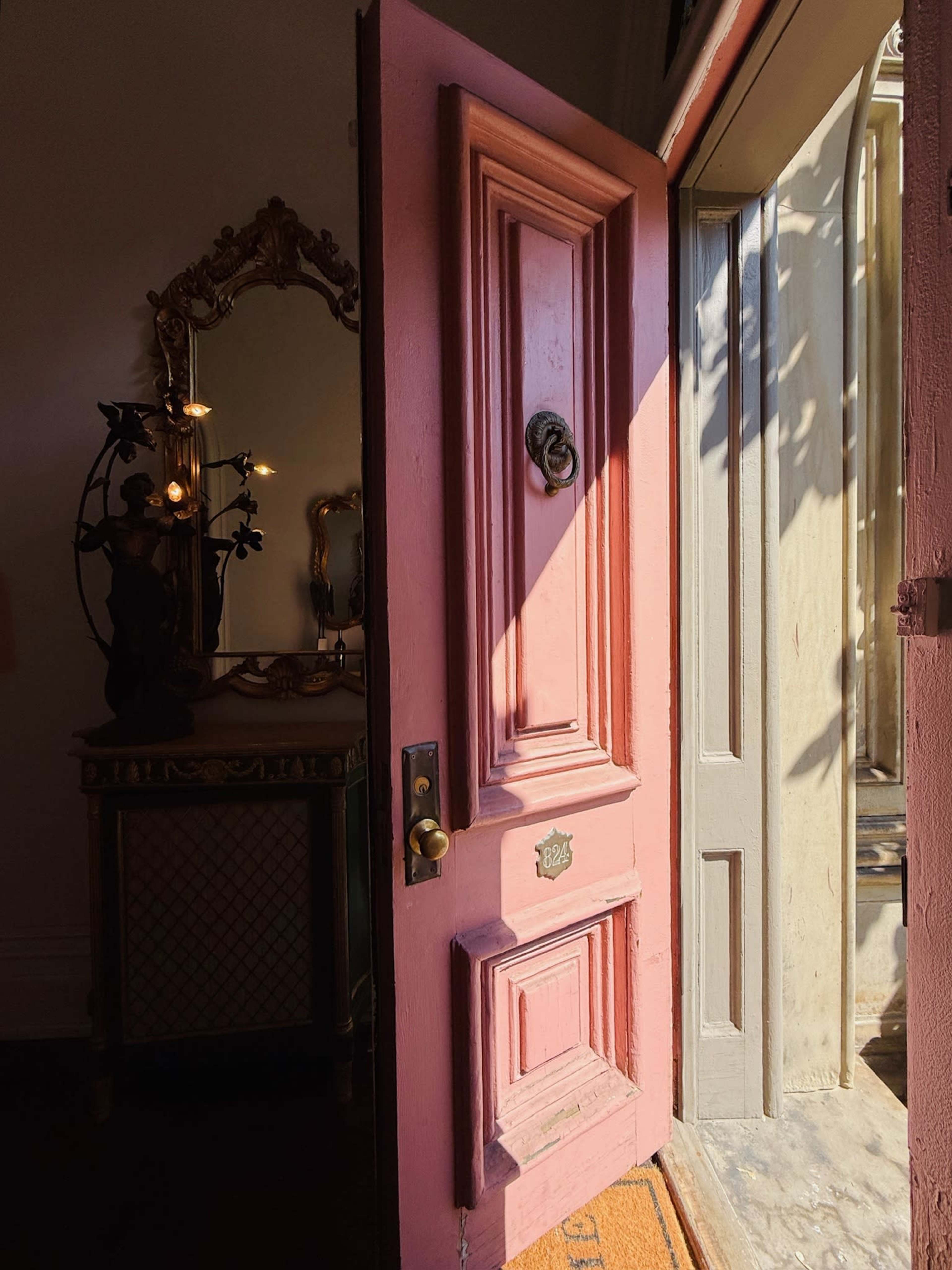 A pink door stands partially open, letting in sunlight and casting shadows on the floor, while a decorative mirror and a statue are visible nearby.