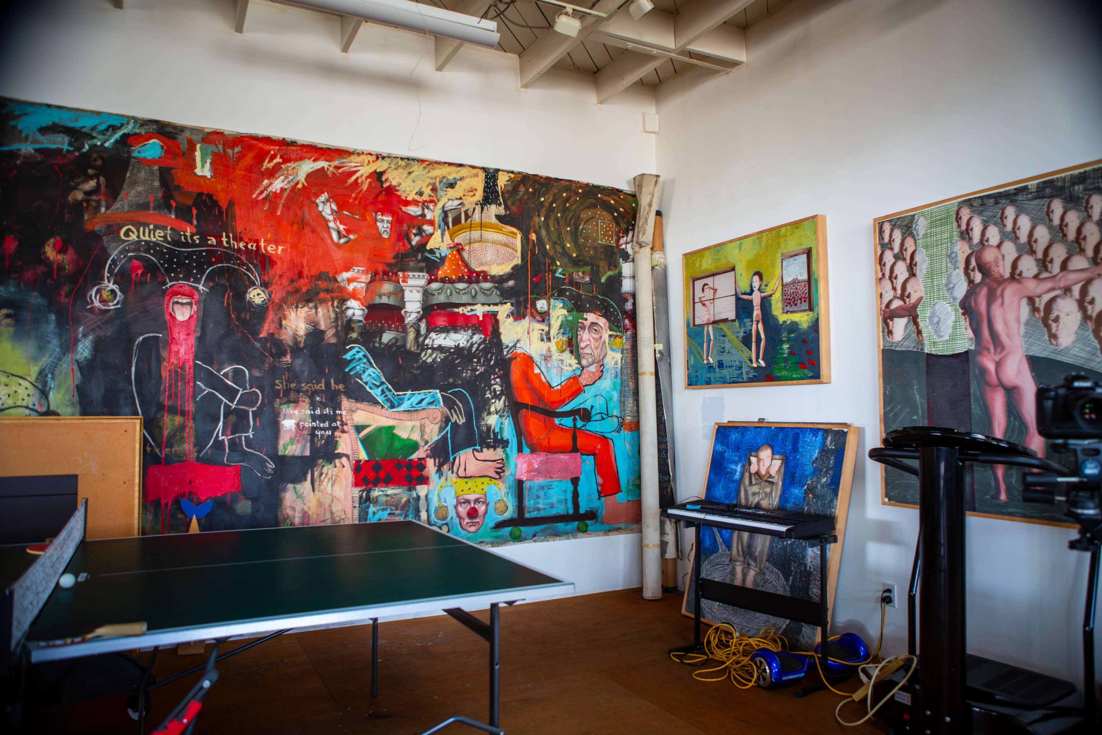 The image shows an art-filled room featuring a ping pong table in the foreground and vibrant wall art displaying various abstract and figurative styles.