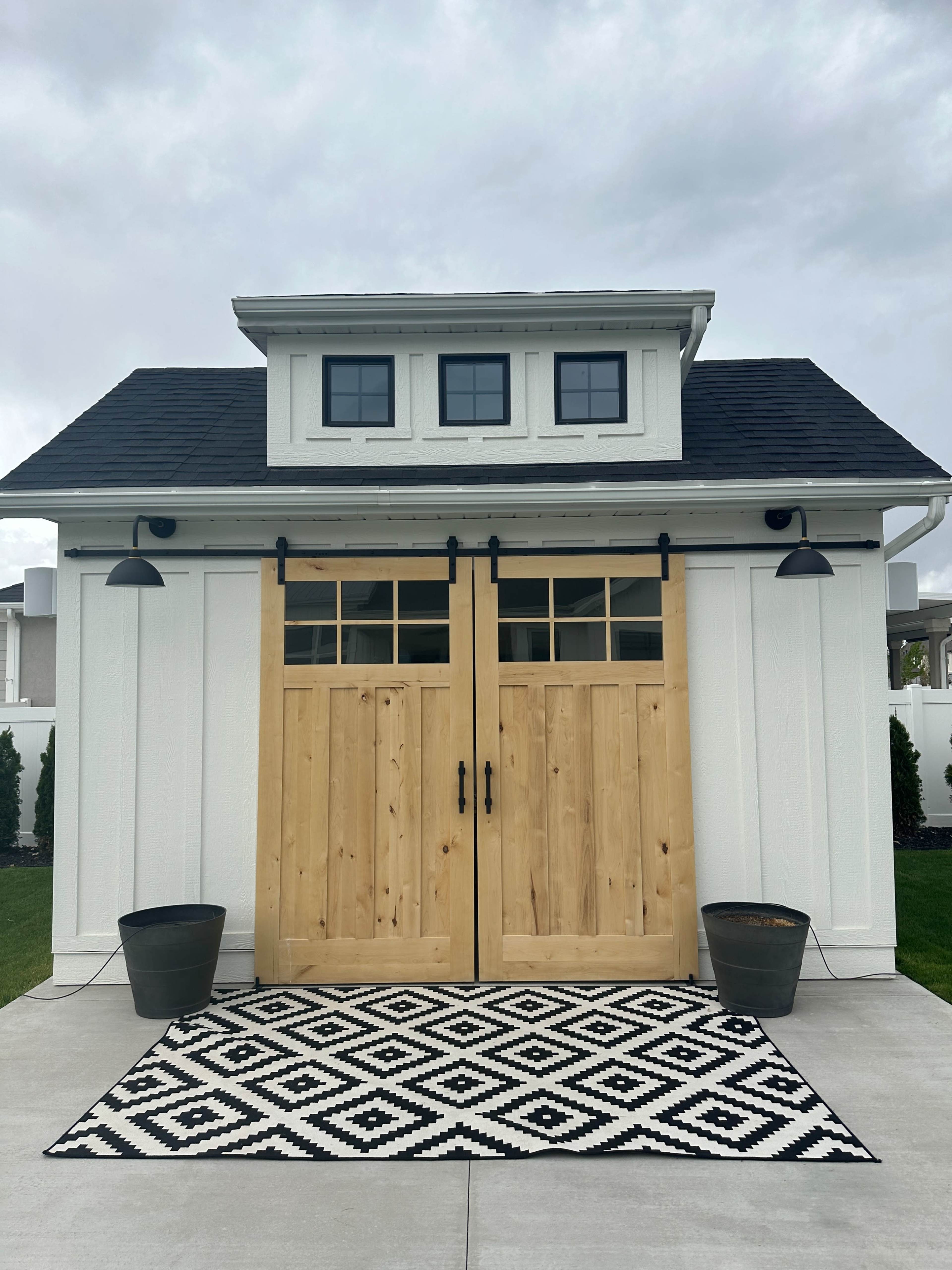 The image shows a white building with large wooden double doors, flanked by two planters on a concrete pathway, under a cloudy sky.