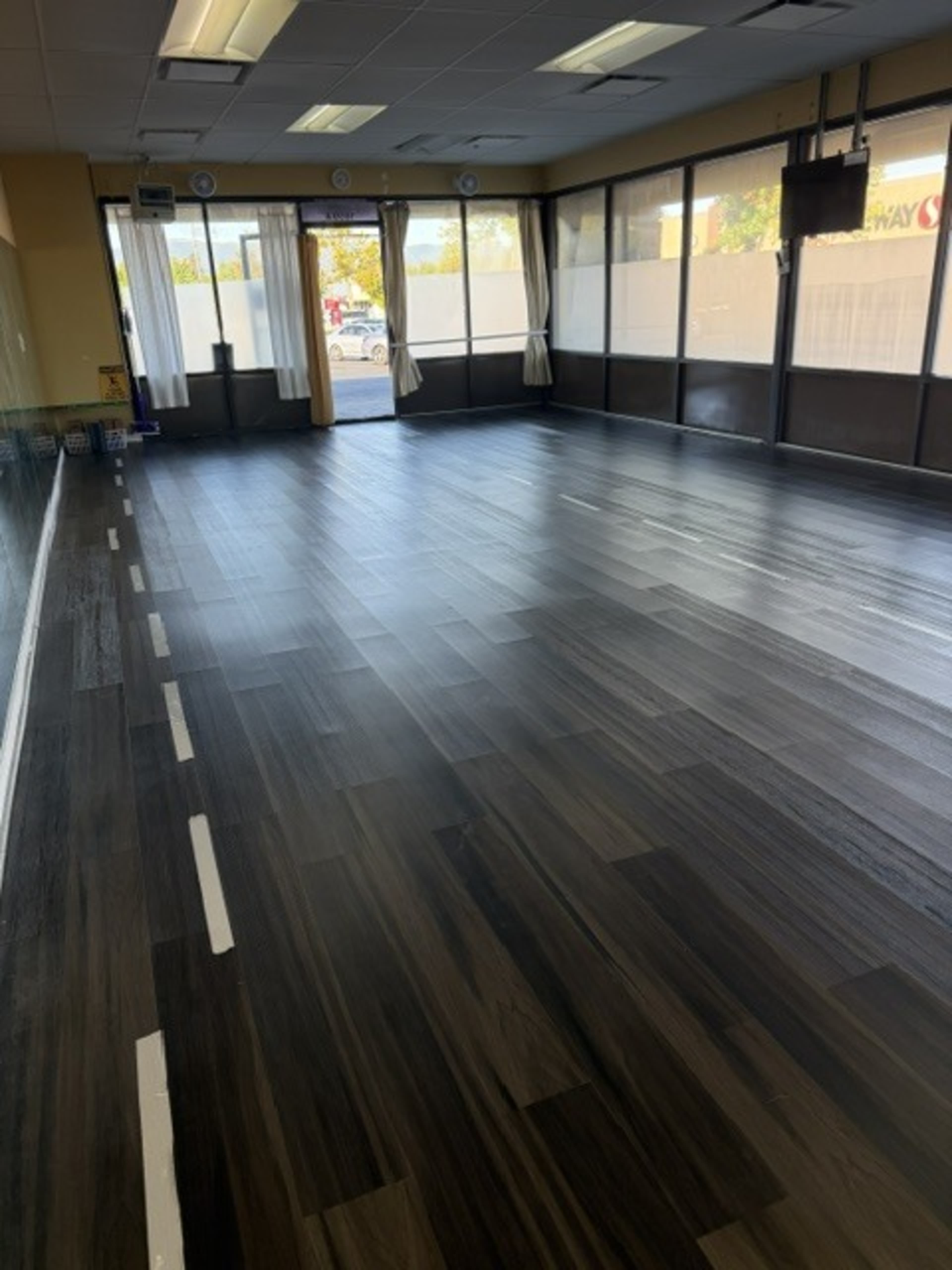 The image shows a spacious room with dark wooden flooring and large windows, partially covered by sheer curtains.