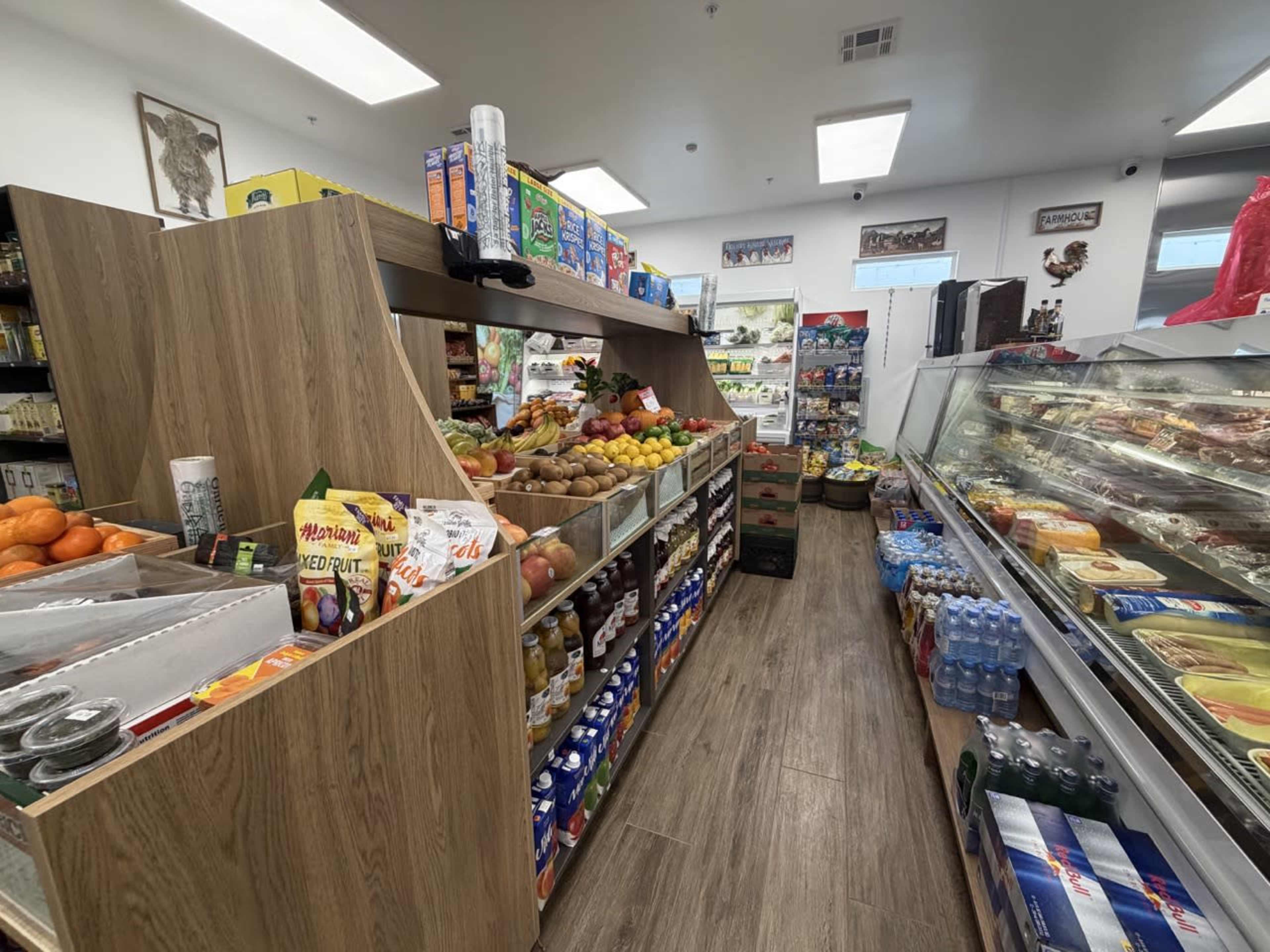 The image shows the interior of a grocery store with wooden shelving displaying various fruits, juices, and snacks, alongside a refrigerated section with packaged food items.