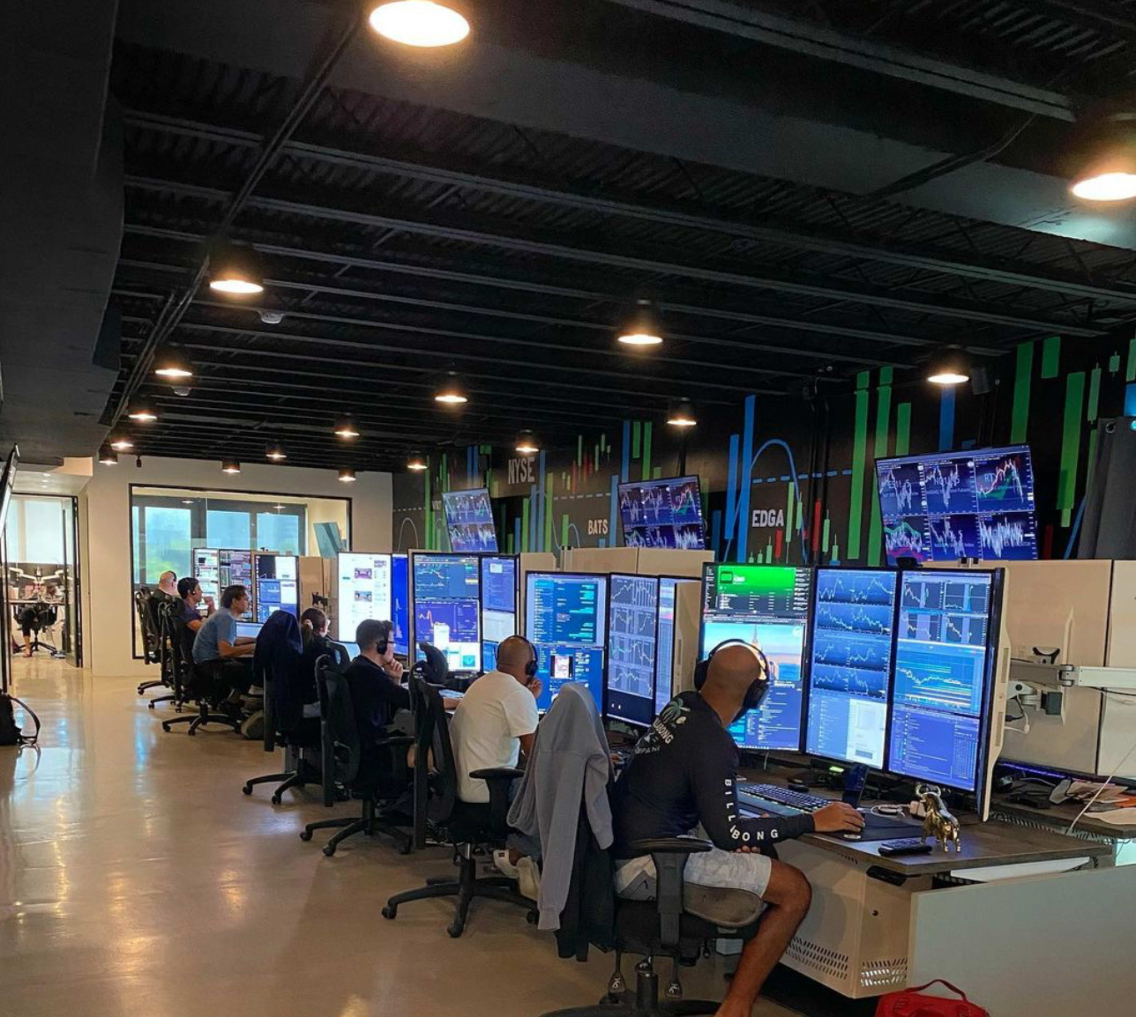 A group of traders works at multiple computer stations in a modern trading office, surrounded by large screens displaying stock market data.