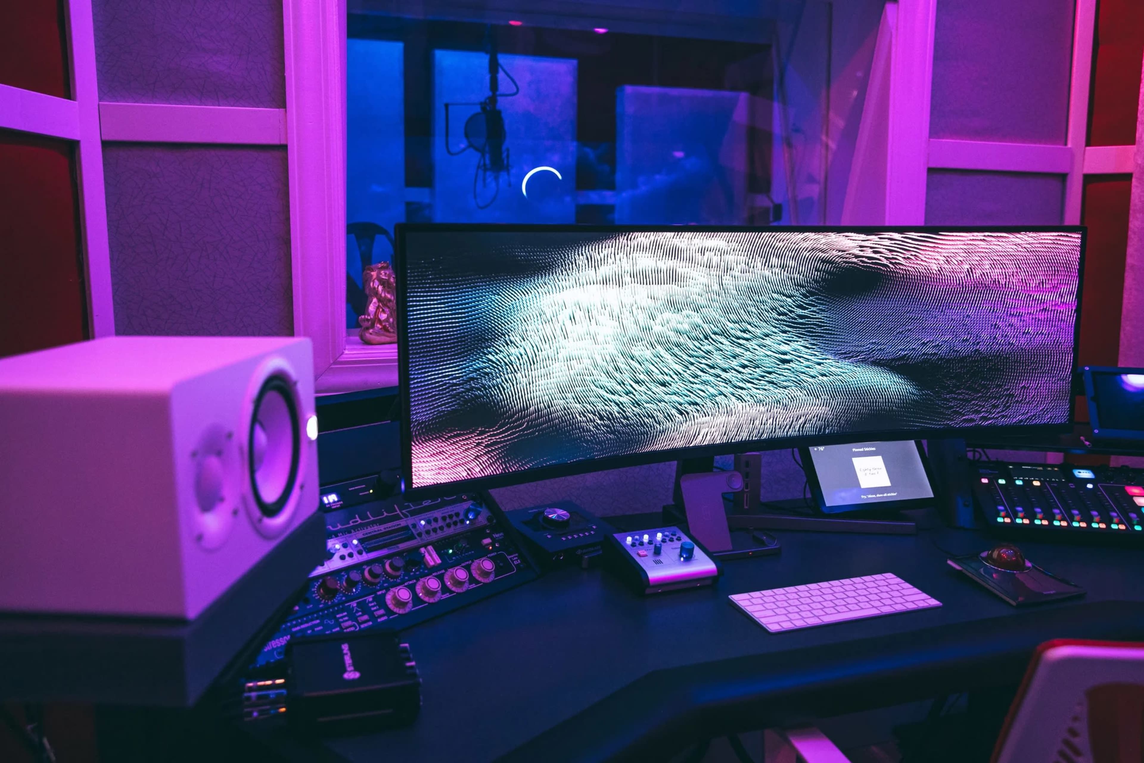 A modern recording studio features a large curved monitor displaying abstract imagery, accompanied by audio equipment and a keyboard on a desk.