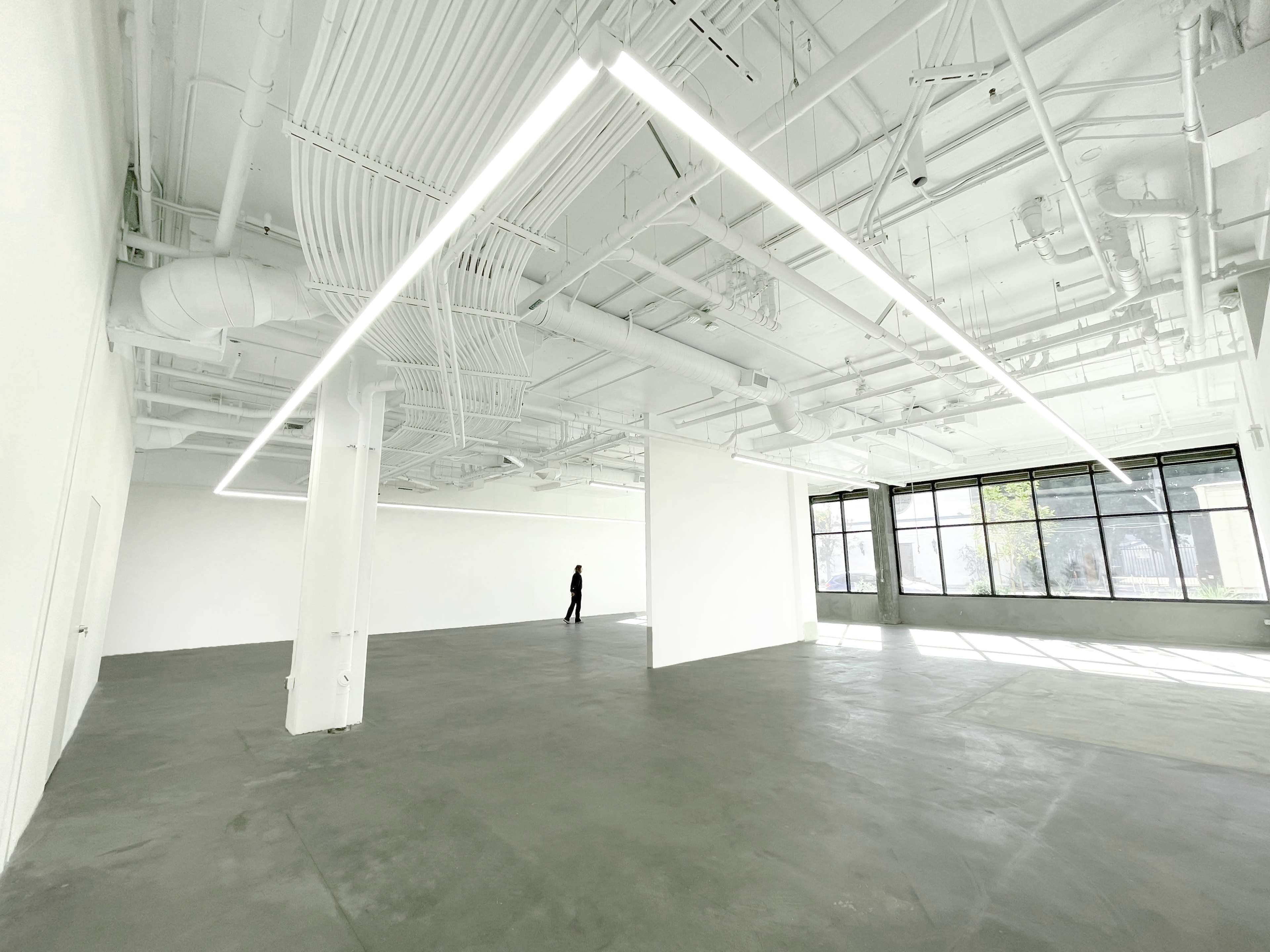 A person stands in an empty, well-lit gallery space with white walls and a concrete floor.