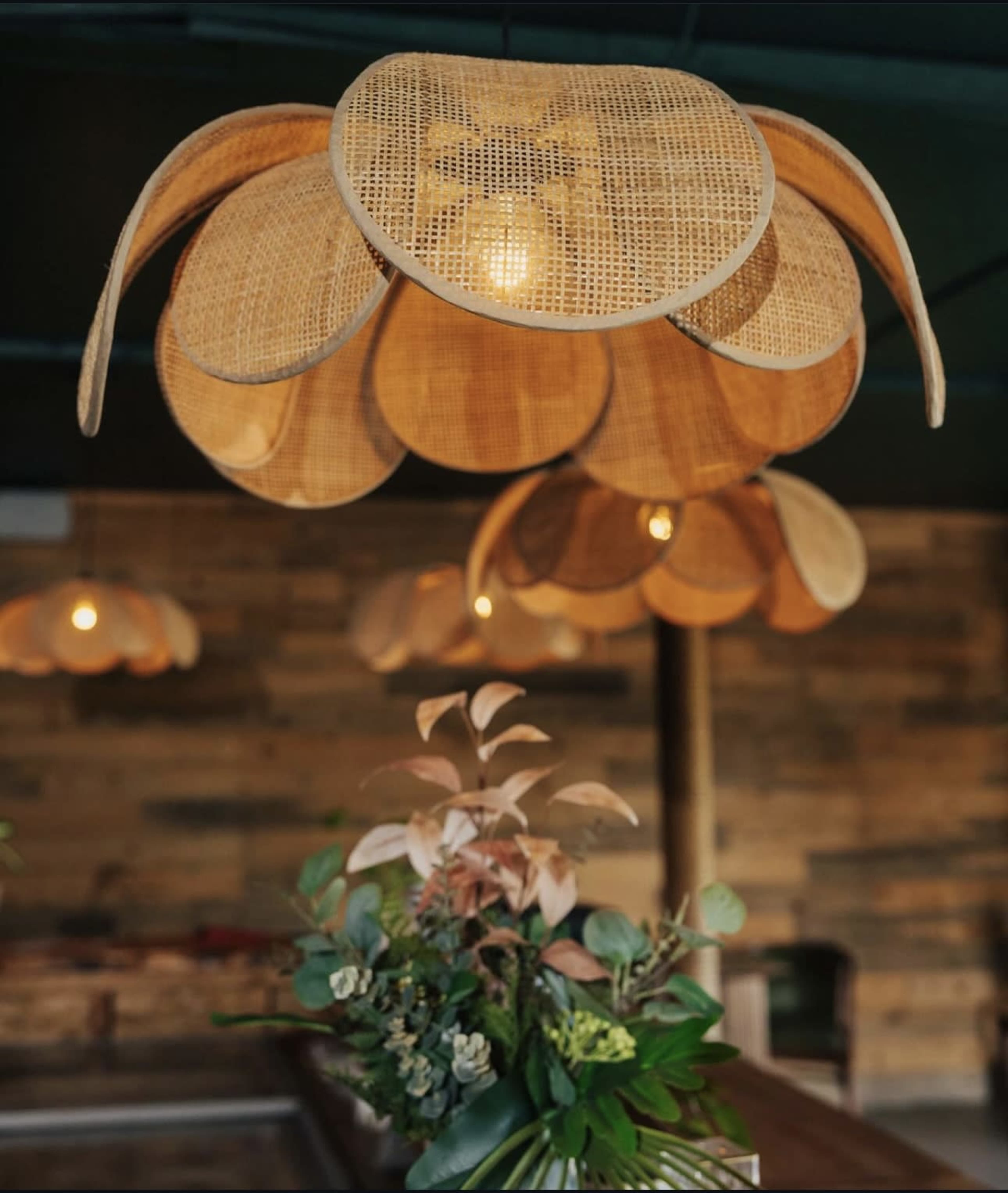 The image depicts woven rattan pendant lights shaped like flowers hanging over a table adorned with greenery and floral arrangements in a rustic wooden interior.