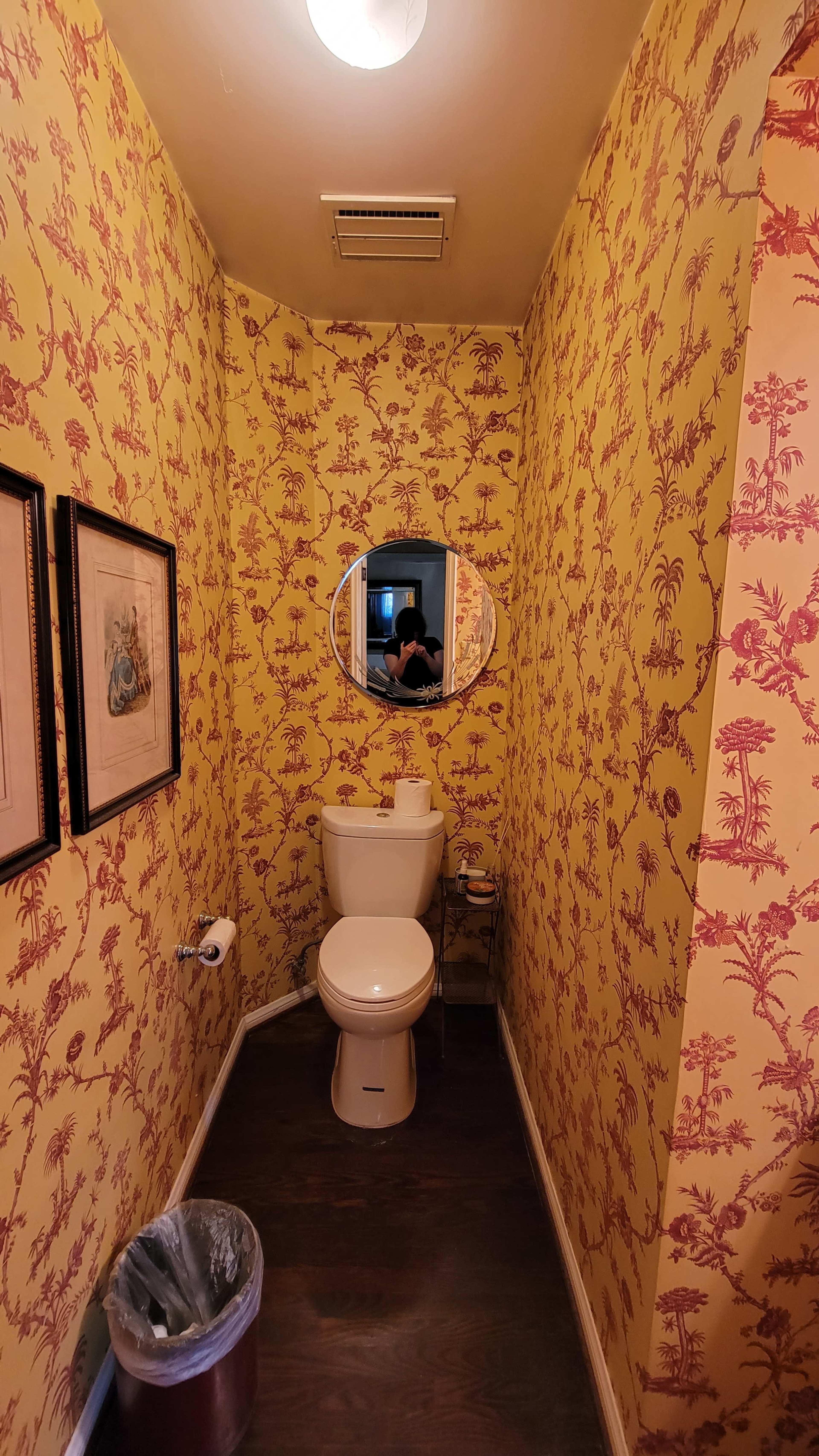 A narrow bathroom with yellow wallpaper featuring a floral pattern, a round mirror above a toilet, and a small trash can on the floor.