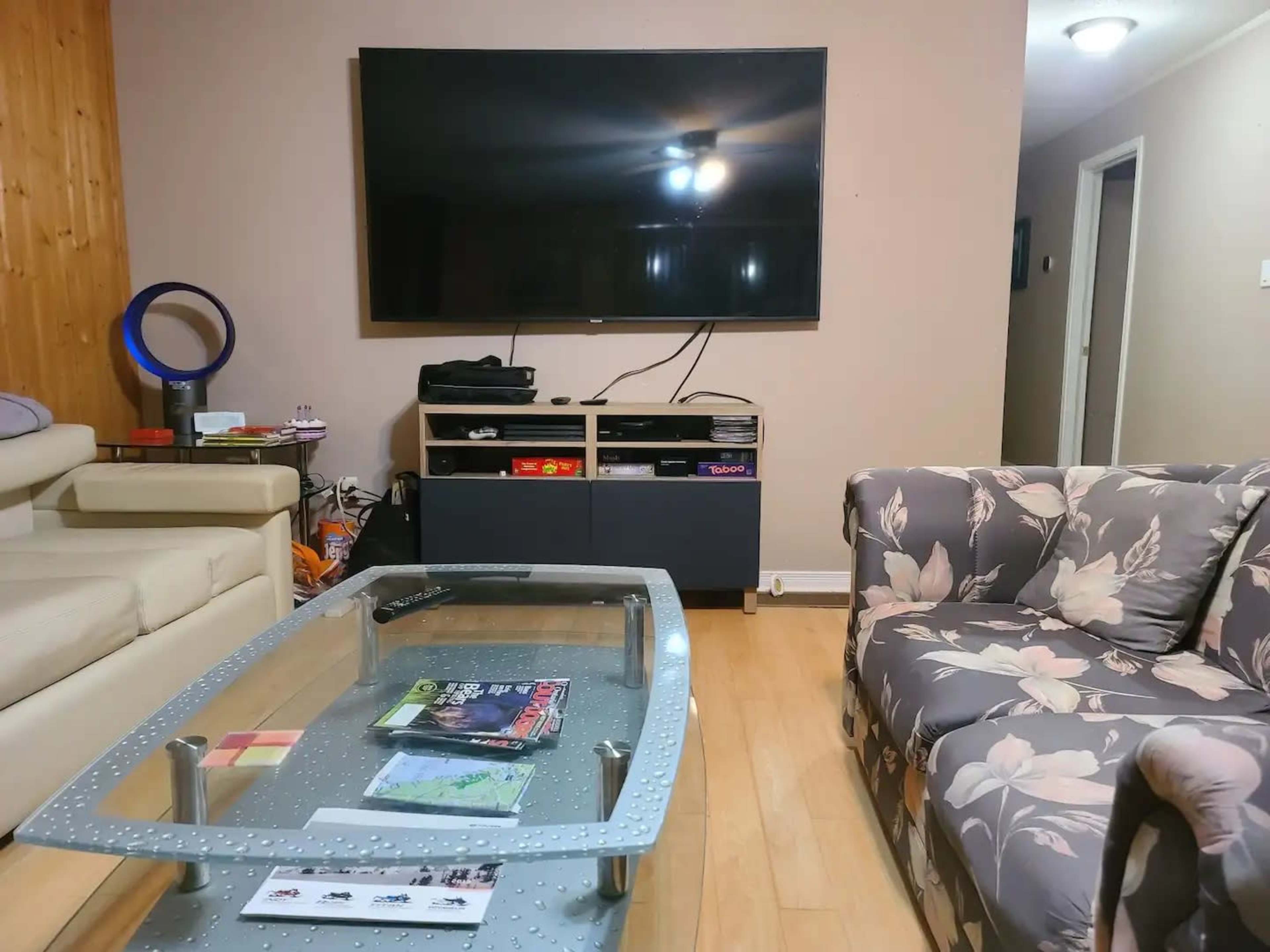A living room features a large flat-screen TV mounted on the wall, a glass coffee table with magazines, and two sofas, one beige and the other with a floral pattern.