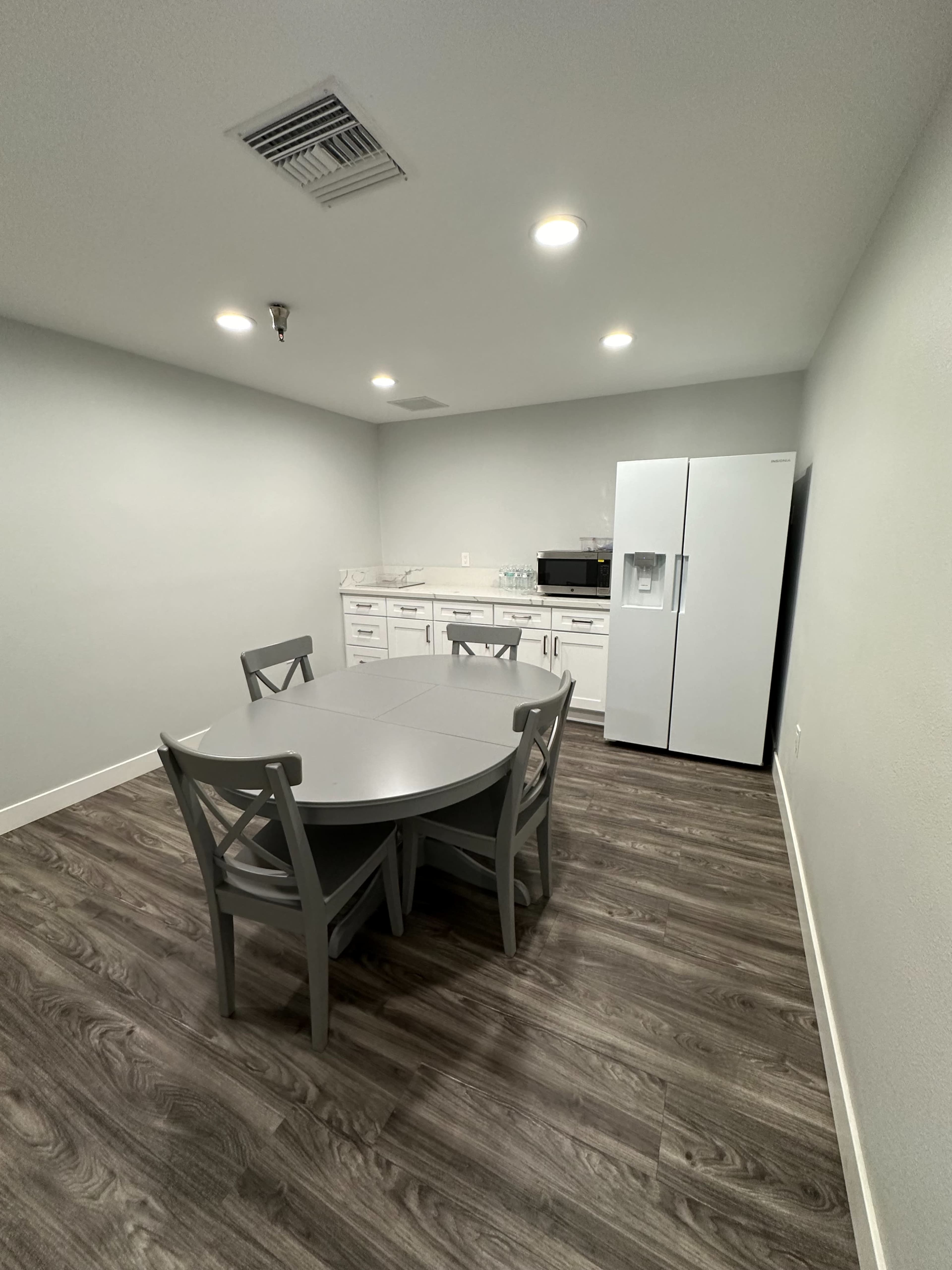 The image shows a kitchenette with a round table and four chairs, a refrigerator, and a microwave against a light-colored wall.