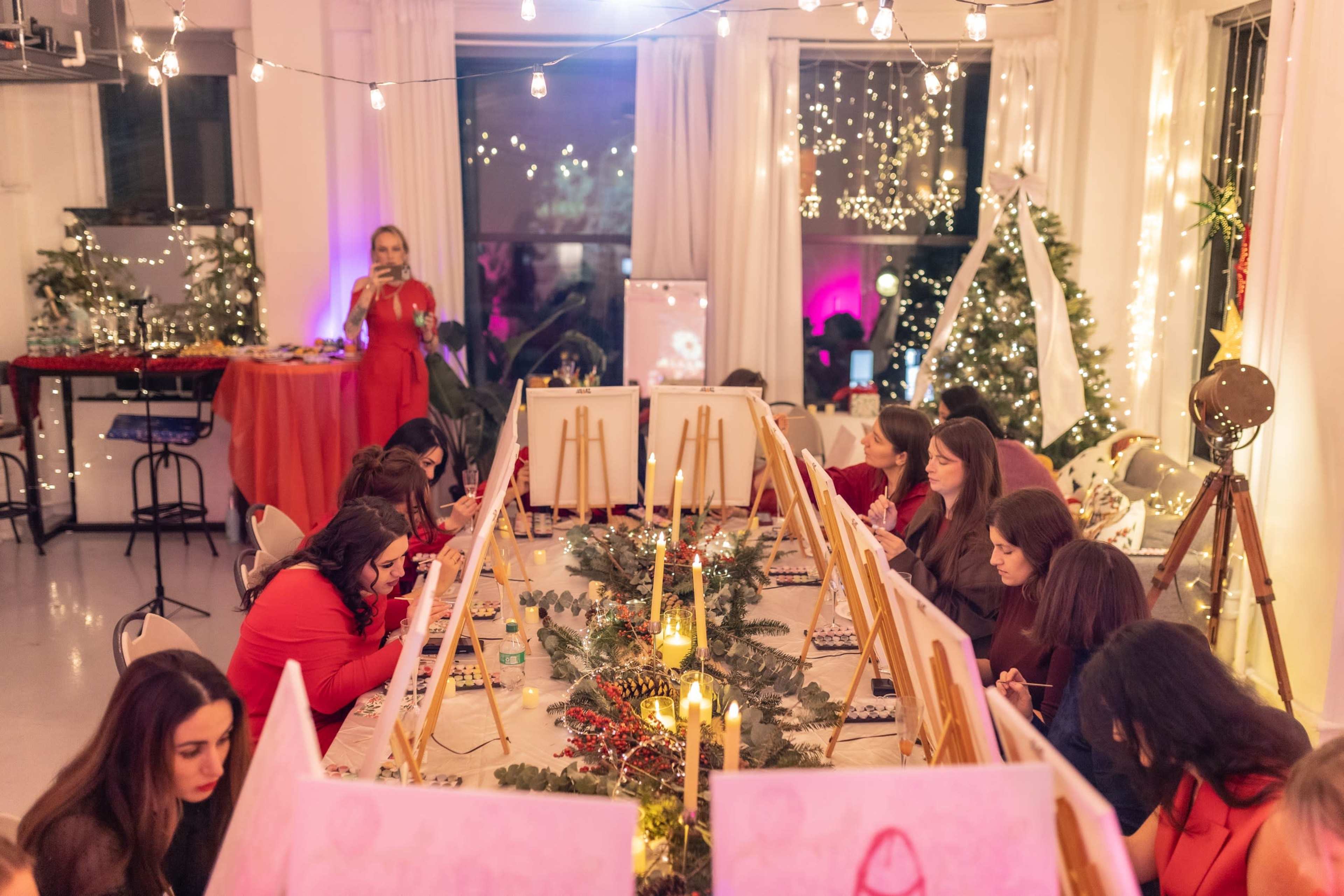A group of women in red attire is seated at a long table, engaged in a painting activity, while the venue is decorated with festive lights and greenery.