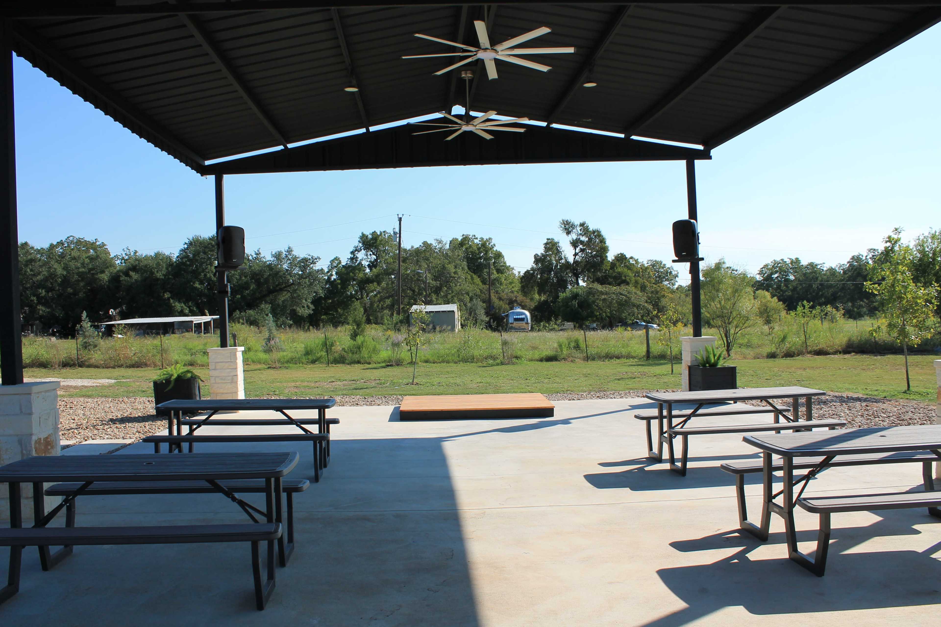The image shows a covered outdoor patio with picnic tables facing a small stage in a grassy area surrounded by trees.