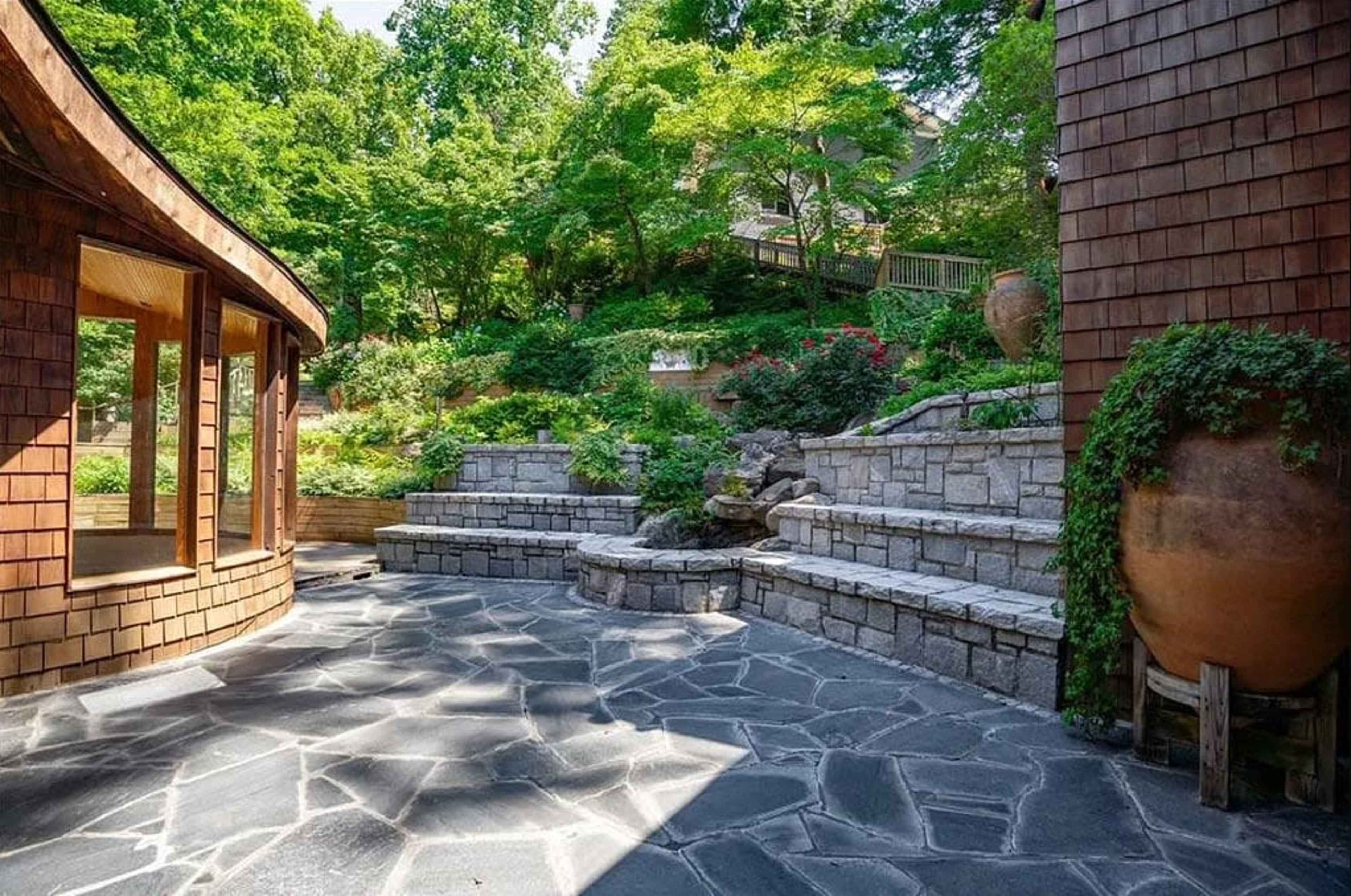 The image shows a landscaped outdoor area featuring stone steps, a patio with a patterned stone floor, and greenery surrounding a water feature.