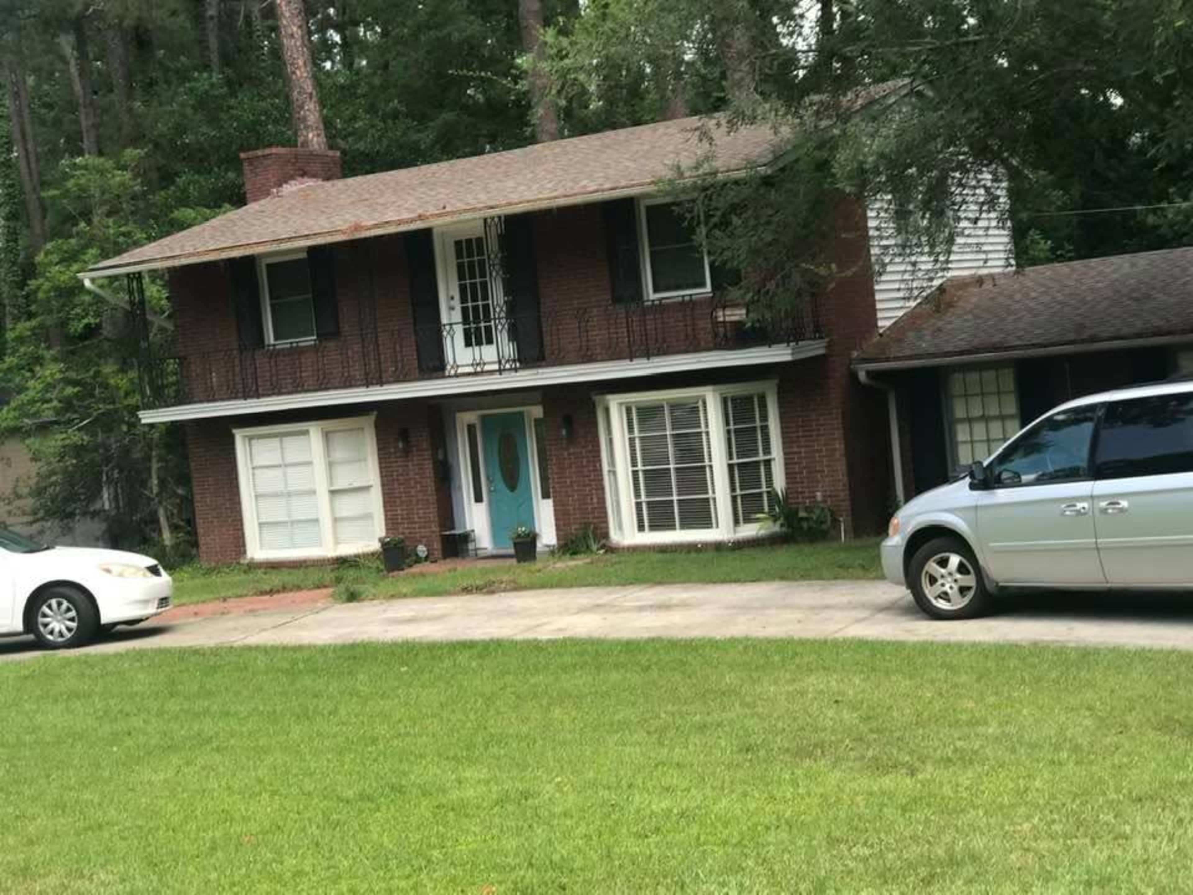 A two-story brick house with a front balcony, surrounded by trees, features a grassy lawn and two parked vehicles in the driveway.