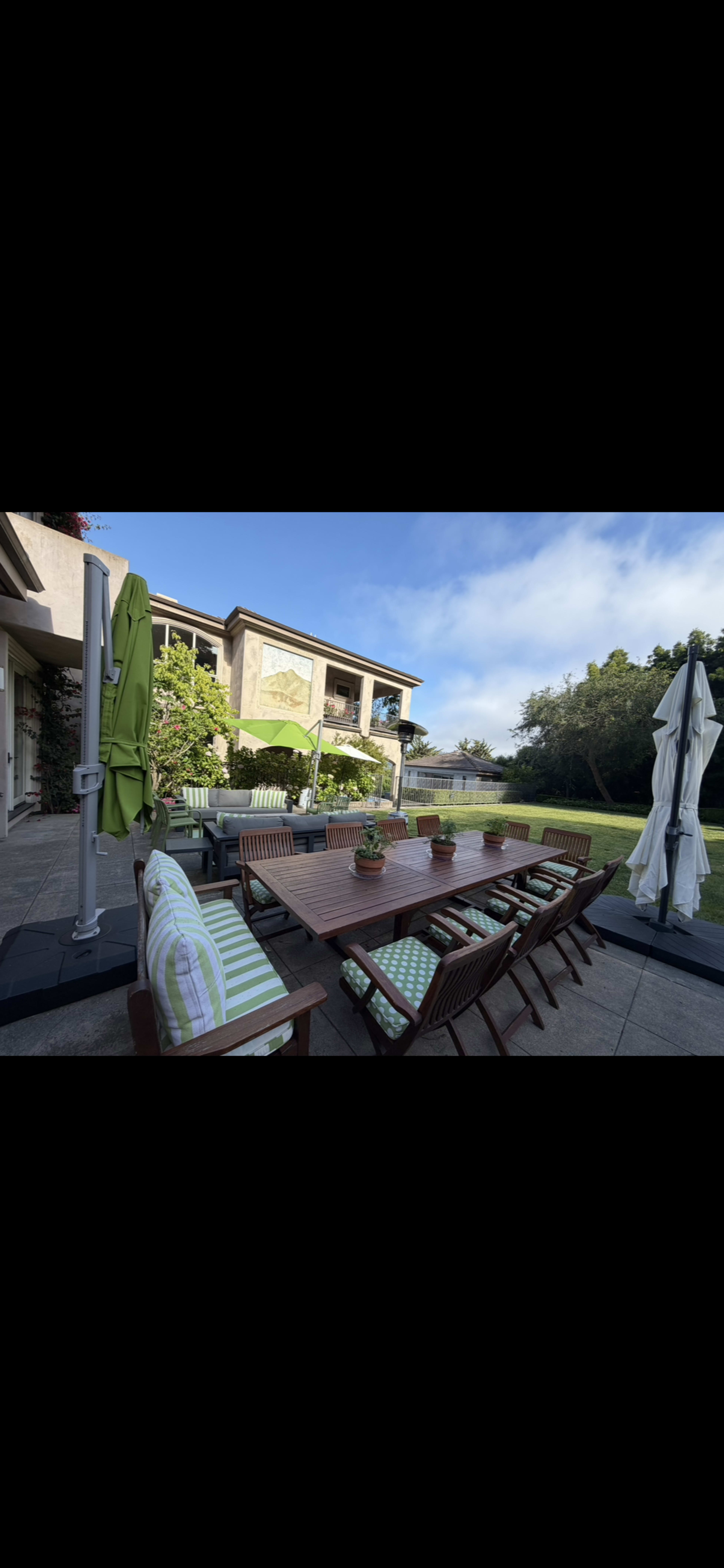 The image shows a well-furnished outdoor dining area with a large wooden table surrounded by matching chairs, set against a backdrop of a modern house and lush greenery.