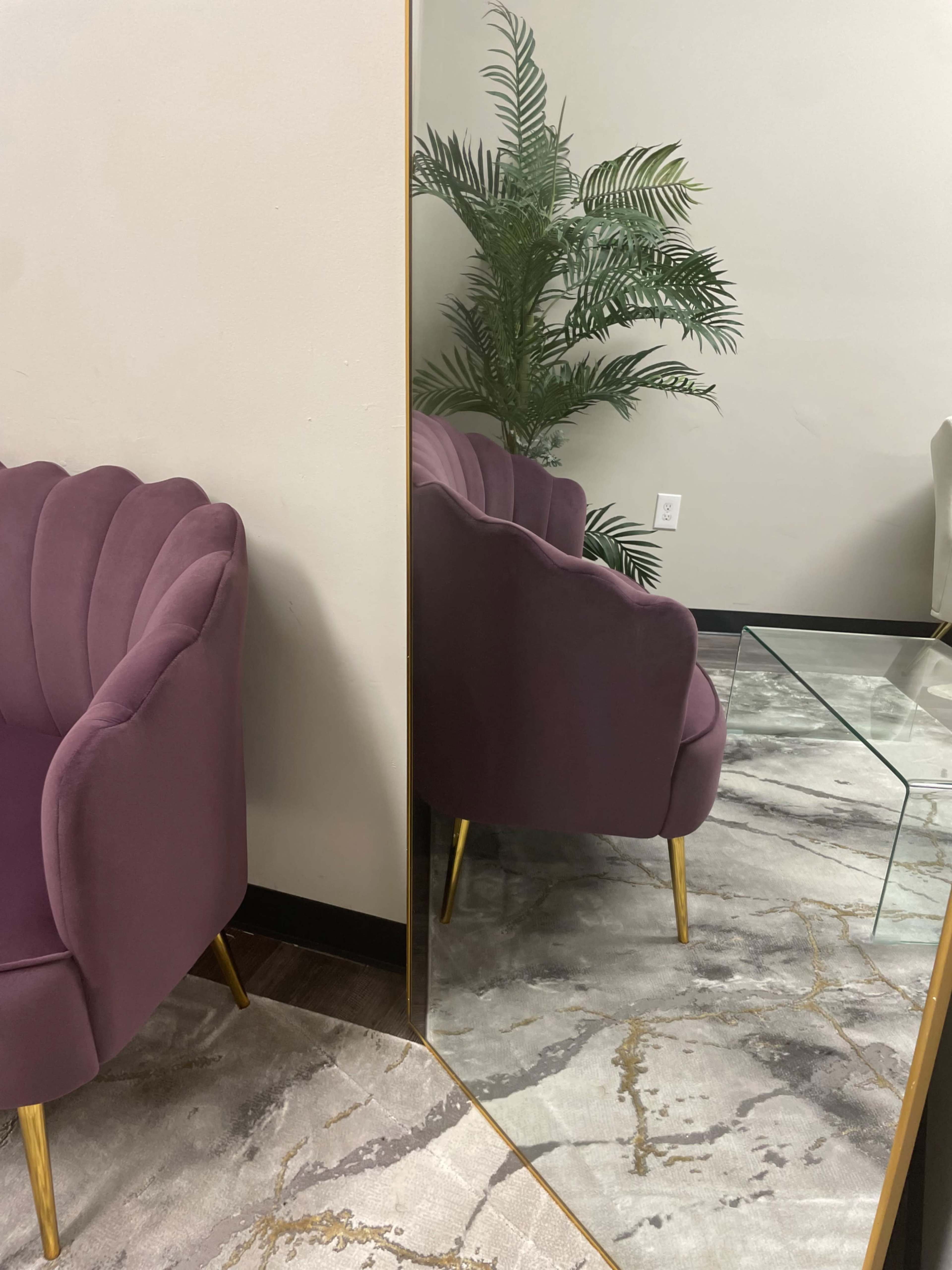 A purple upholstered chair is positioned near a mirror, reflecting a potted plant and a glass table on a marbled floor.