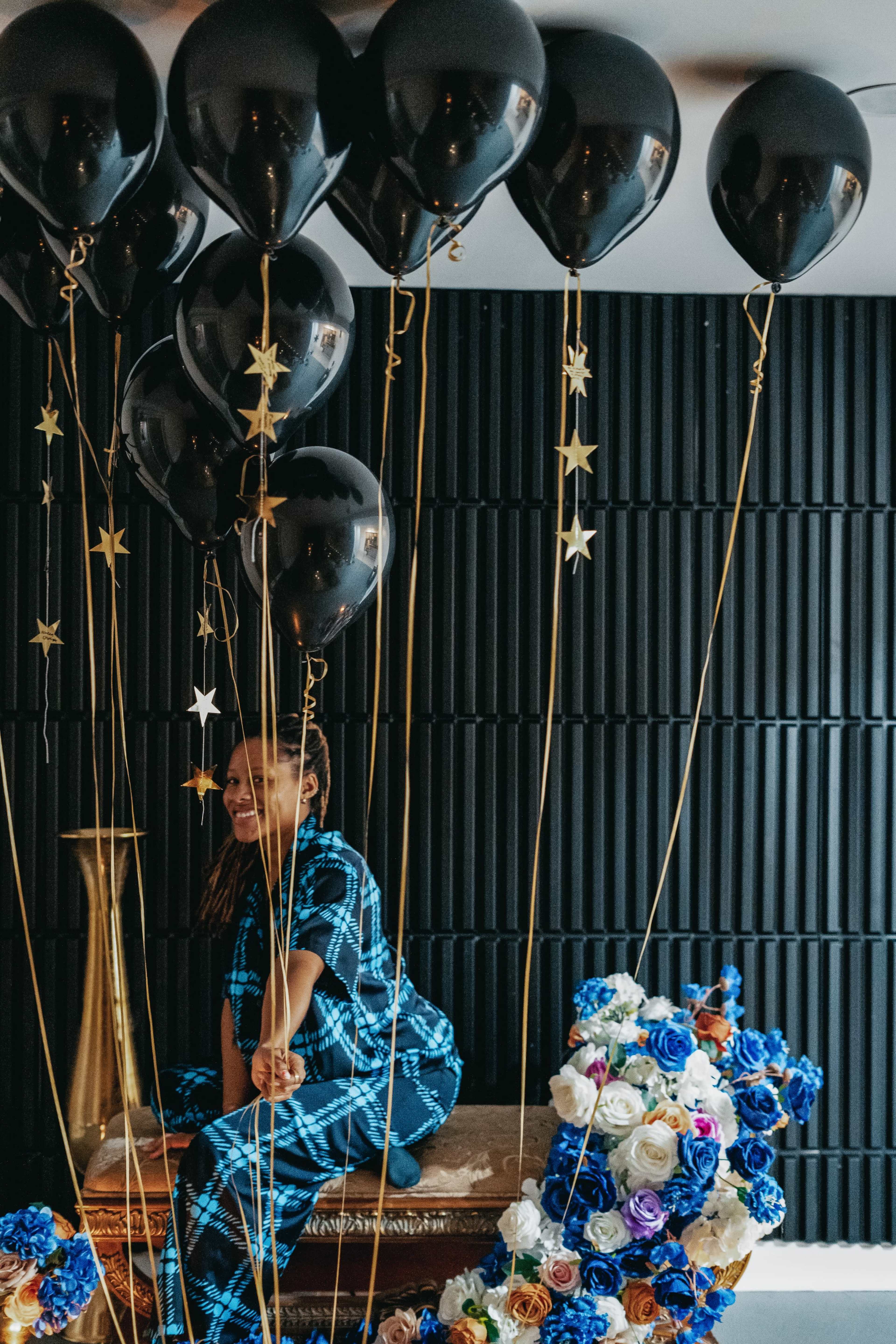 A person sits on a decorative table surrounded by black balloons and a floral arrangement of blue and white roses.