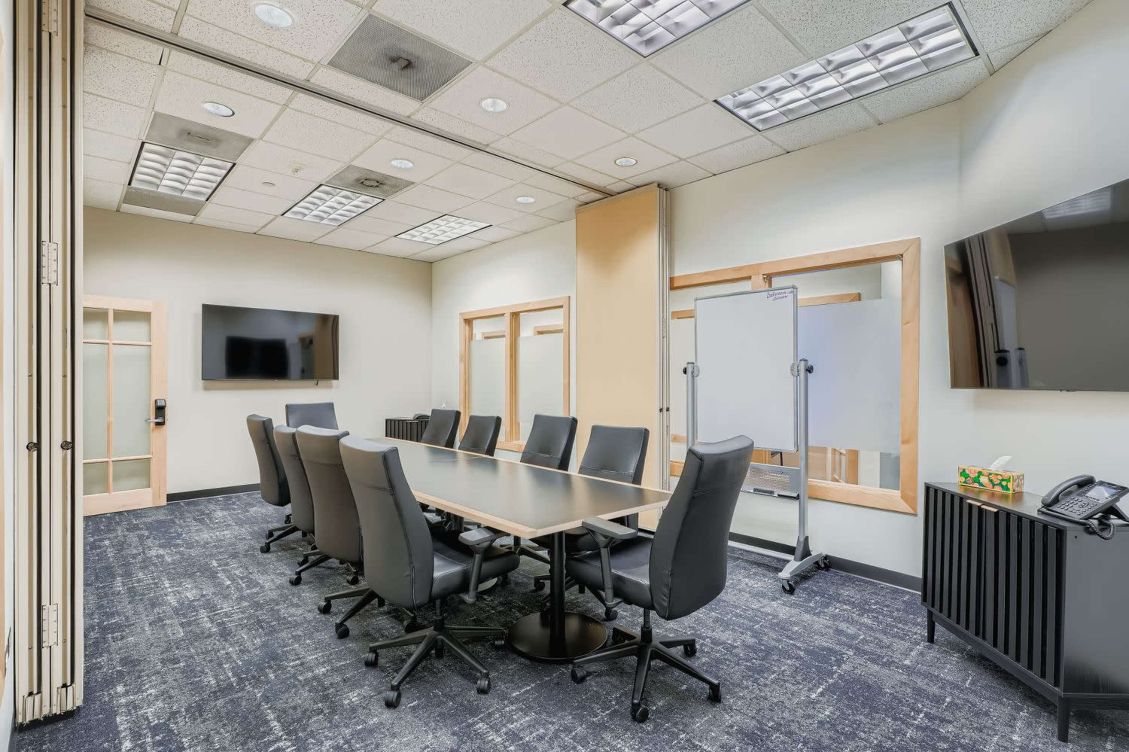 The image shows a conference room with a long table surrounded by black chairs, two wall-mounted televisions, and a whiteboard on a stand.
