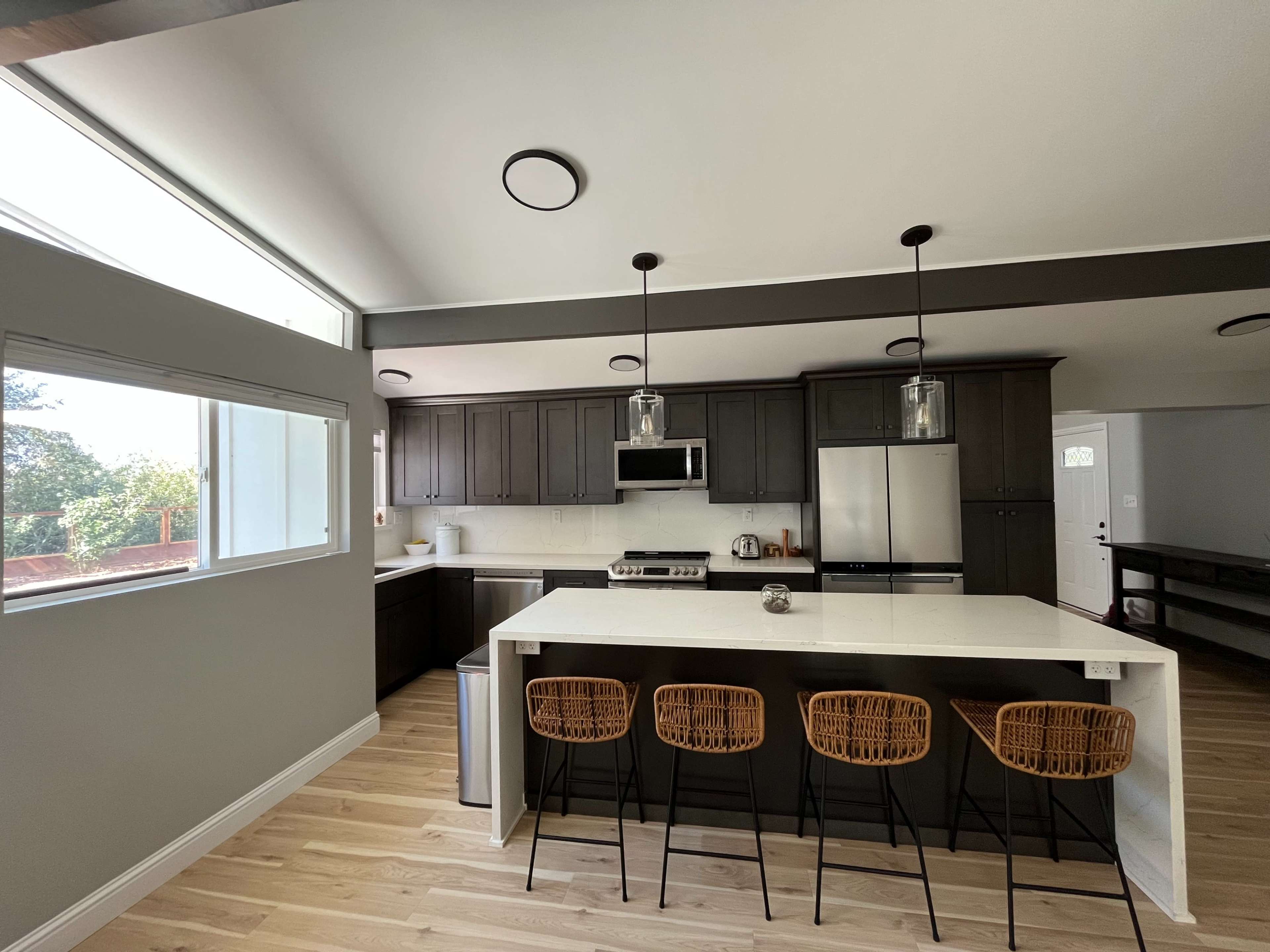A modern kitchen featuring dark cabinets, stainless steel appliances, and a white island with four rattan barstools.