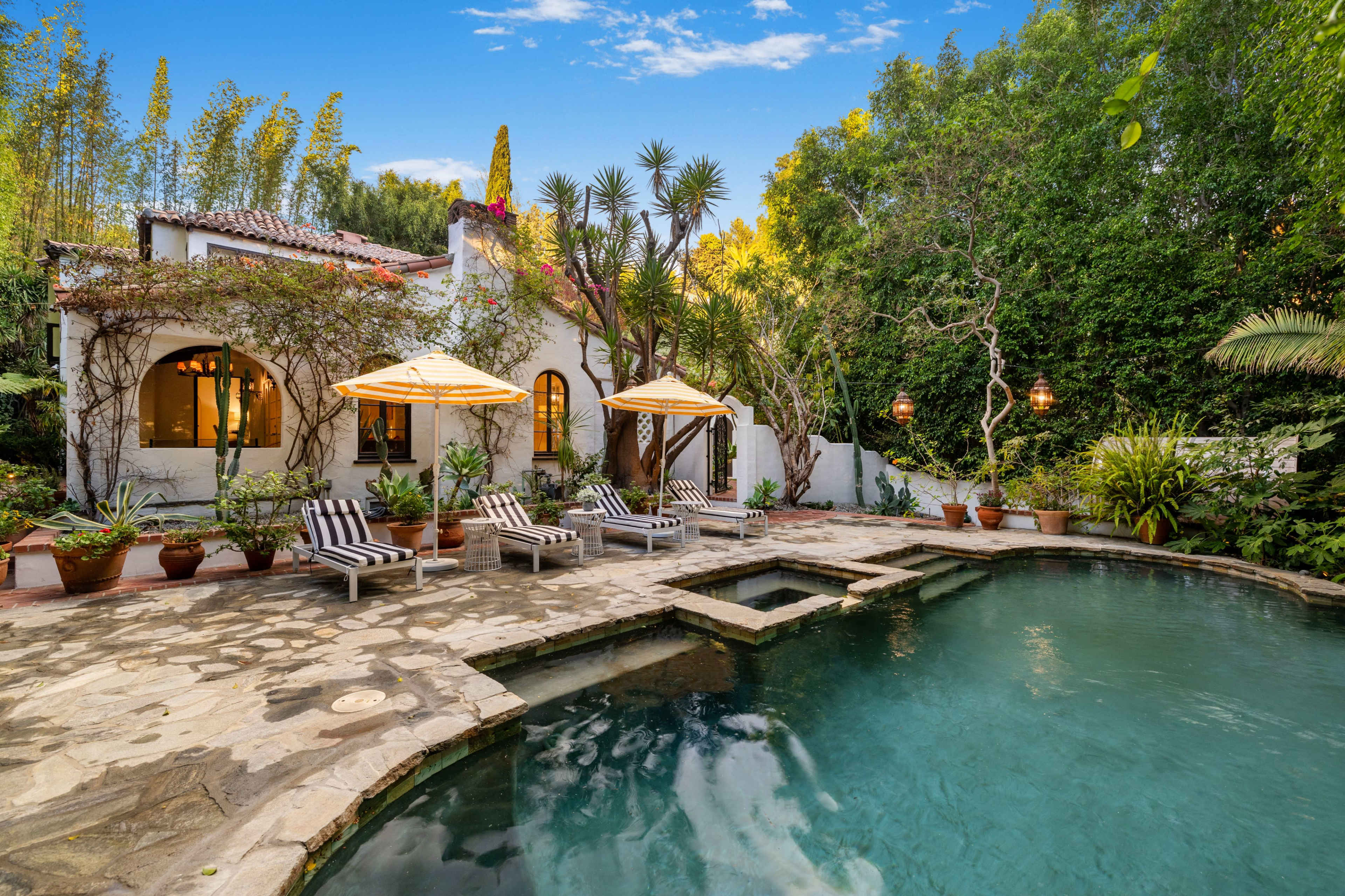 The image features a serene outdoor pool area surrounded by lush greenery and a charming white house with a tiled roof, complemented by lounge chairs and umbrellas.