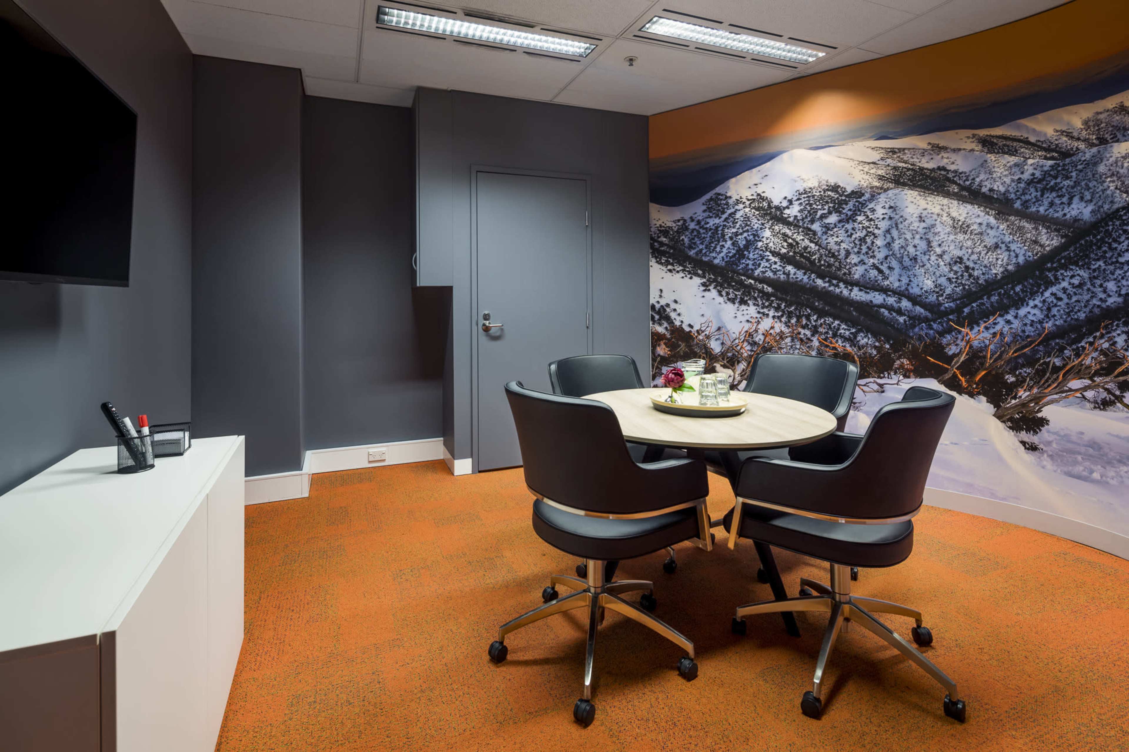 A conference room features a circular table surrounded by four black chairs, with a wall mural depicting a snowy mountain landscape and an orange carpet.