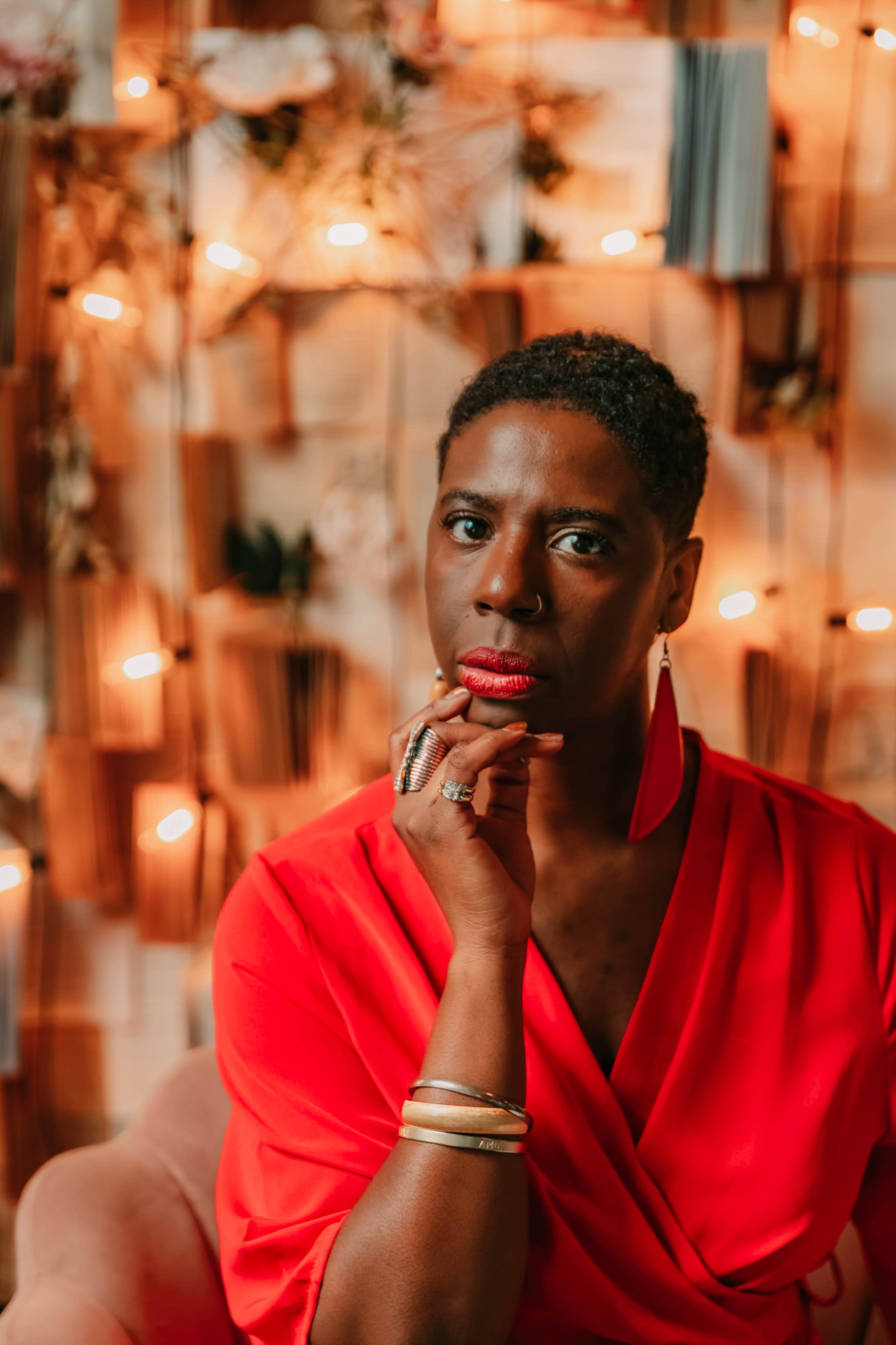 A woman in a red blouse sits thoughtfully with her hand on her chin, against a backdrop of lights and decorative elements.