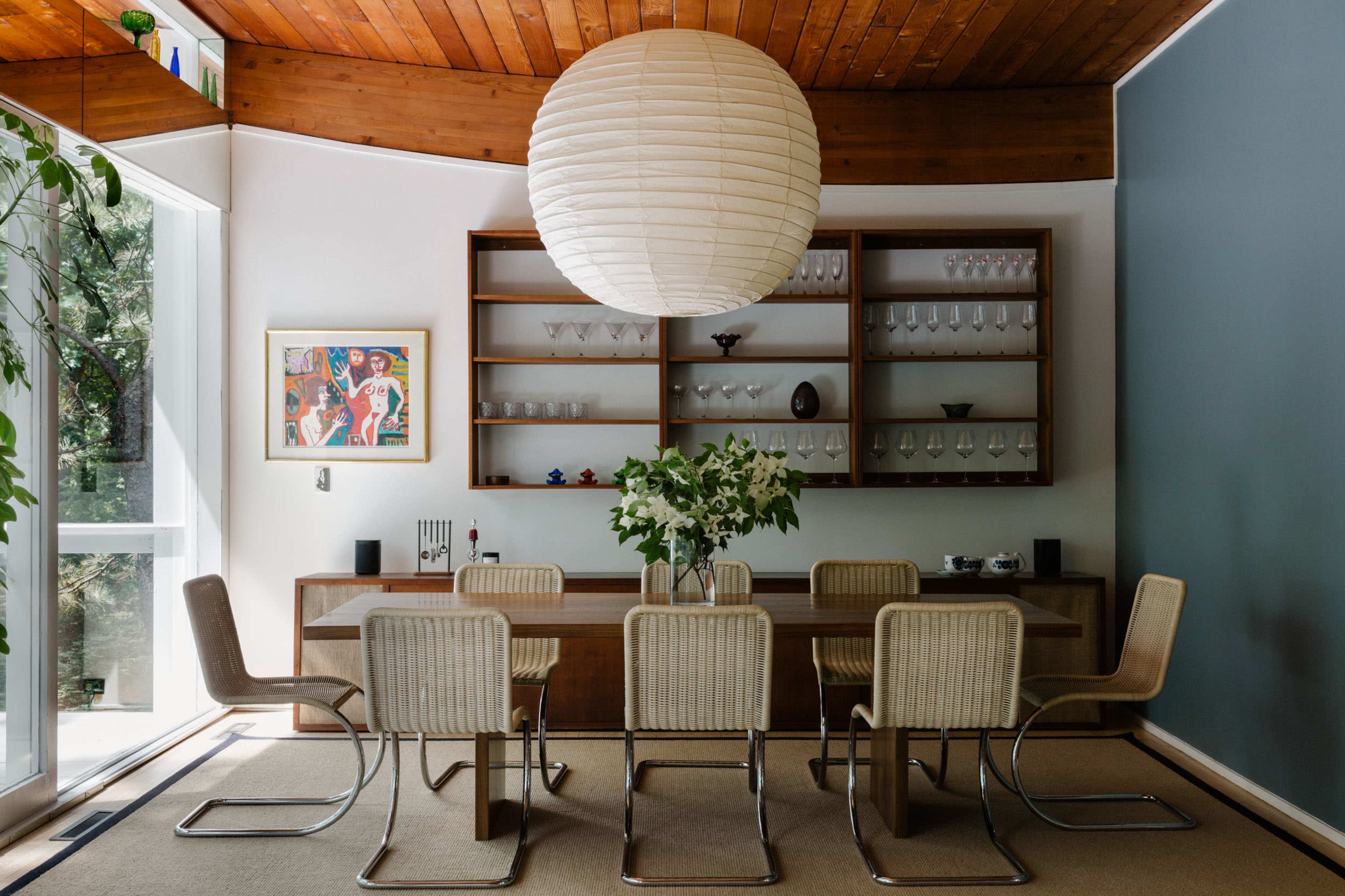 A modern dining room features a large wooden table surrounded by six metal-framed chairs, illuminated by a round paper pendant light, with glassware displayed on shelves above.