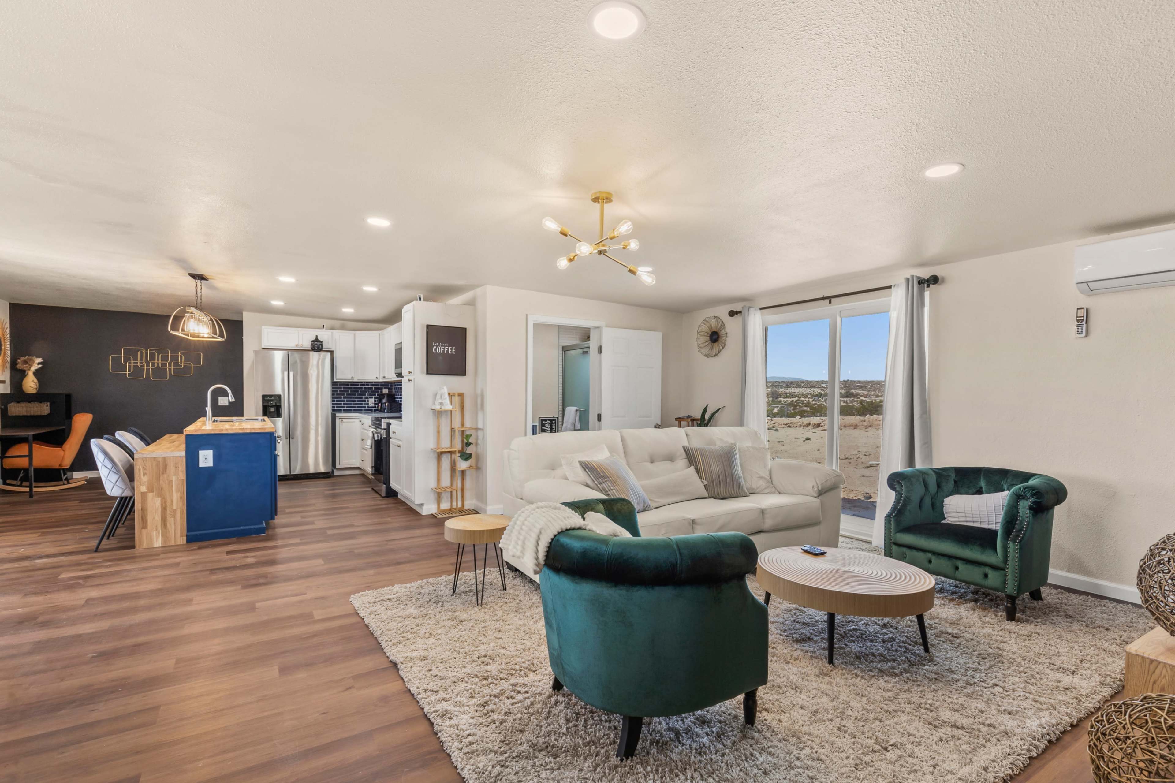 A modern living room features a white sofa, green accent chairs, a coffee table, and an open kitchen area with wooden and white cabinetry.