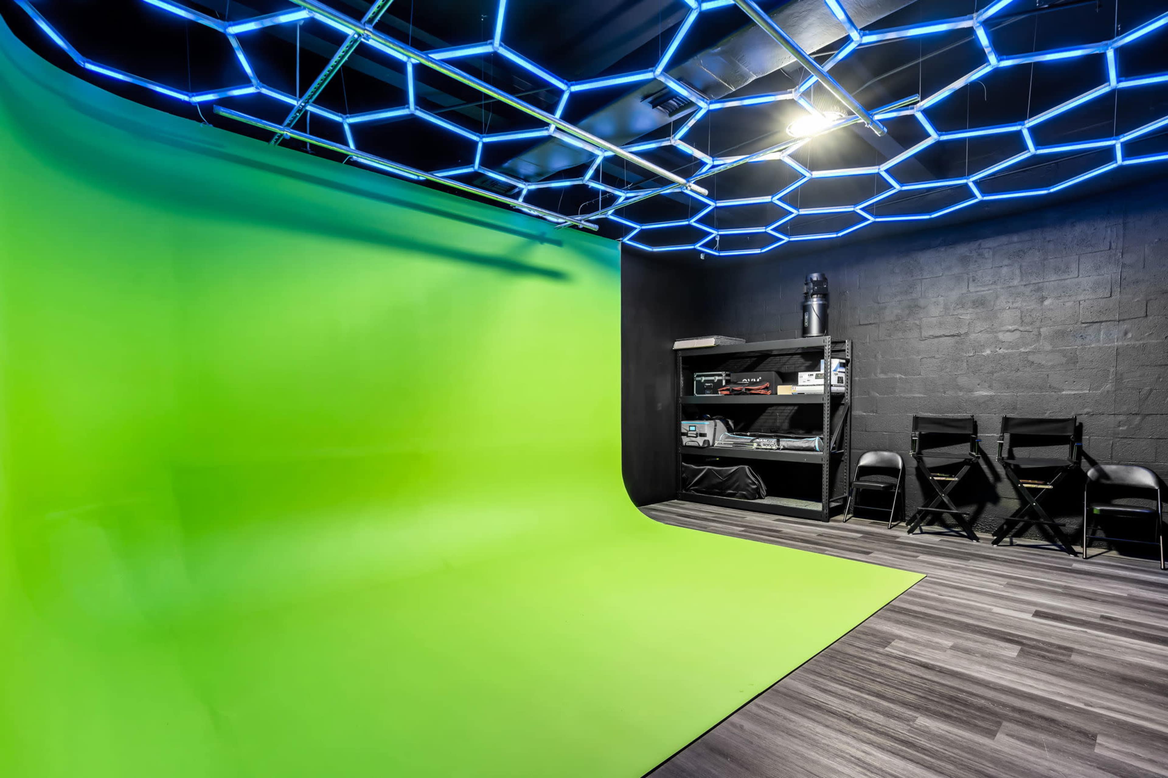 The image shows a green screen backdrop in a studio setting with black walls, a shelf with equipment, and blue LED lighting along the ceiling.