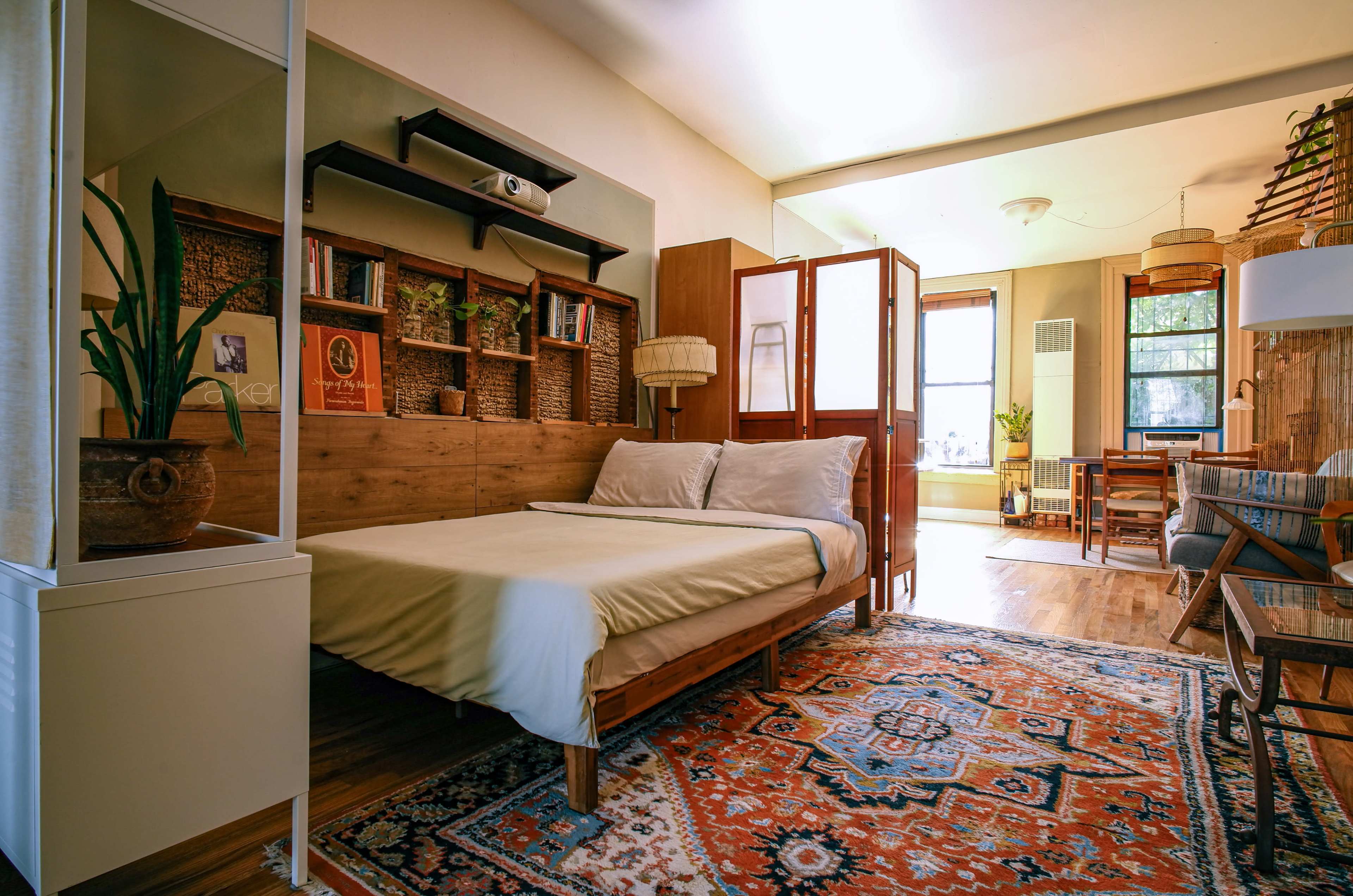 The image shows a cozy bedroom featuring a bed with a simple headboard, a decorative rug, and various furniture pieces including a wooden table and plants.