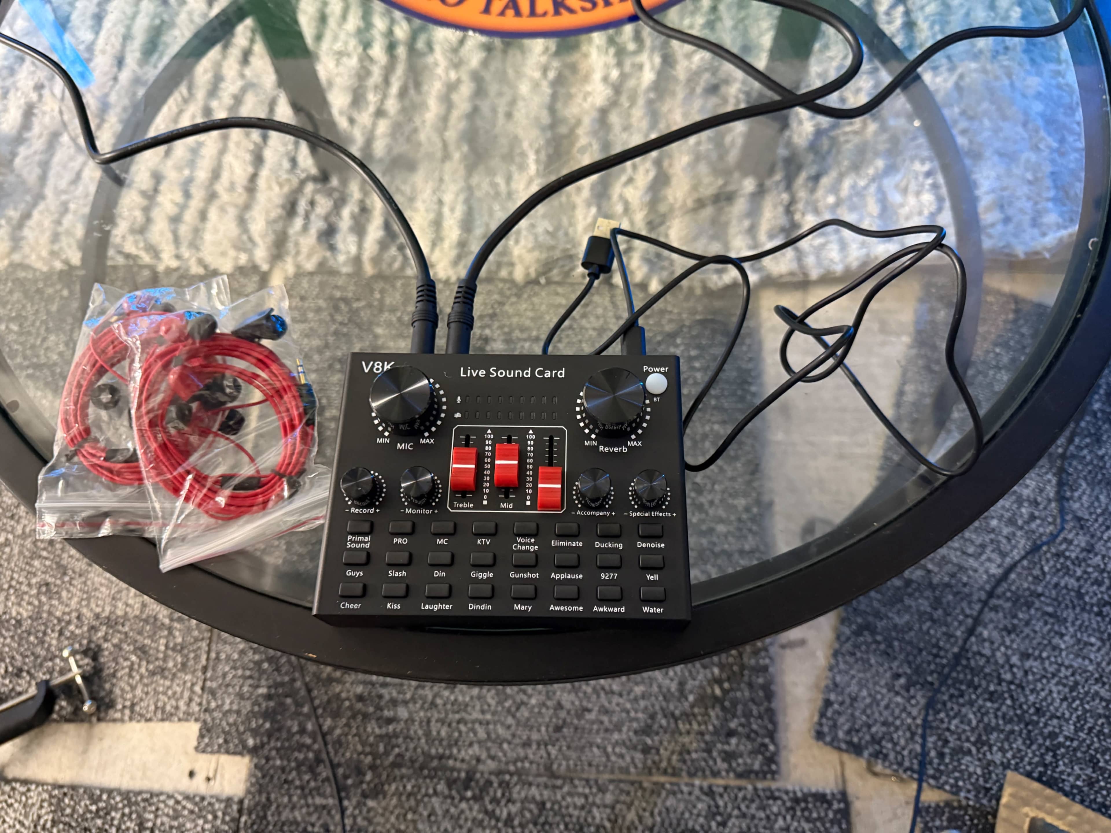 A live sound card with multiple controls and connections is placed on a glass table next to two packages of red audio cables.