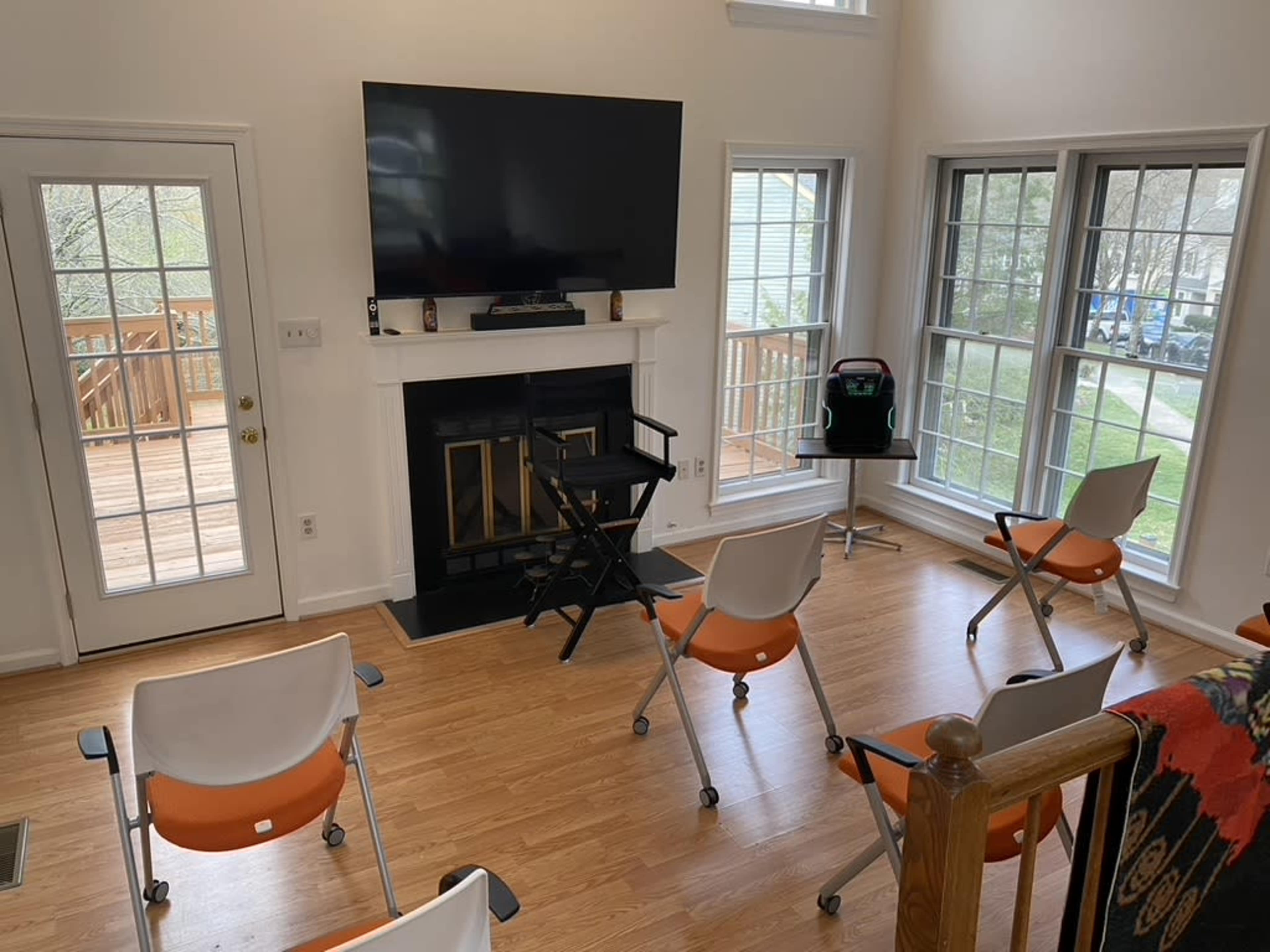 The room features a set of orange rolling chairs arranged facing a mounted television and a fireplace, with large windows providing natural light.