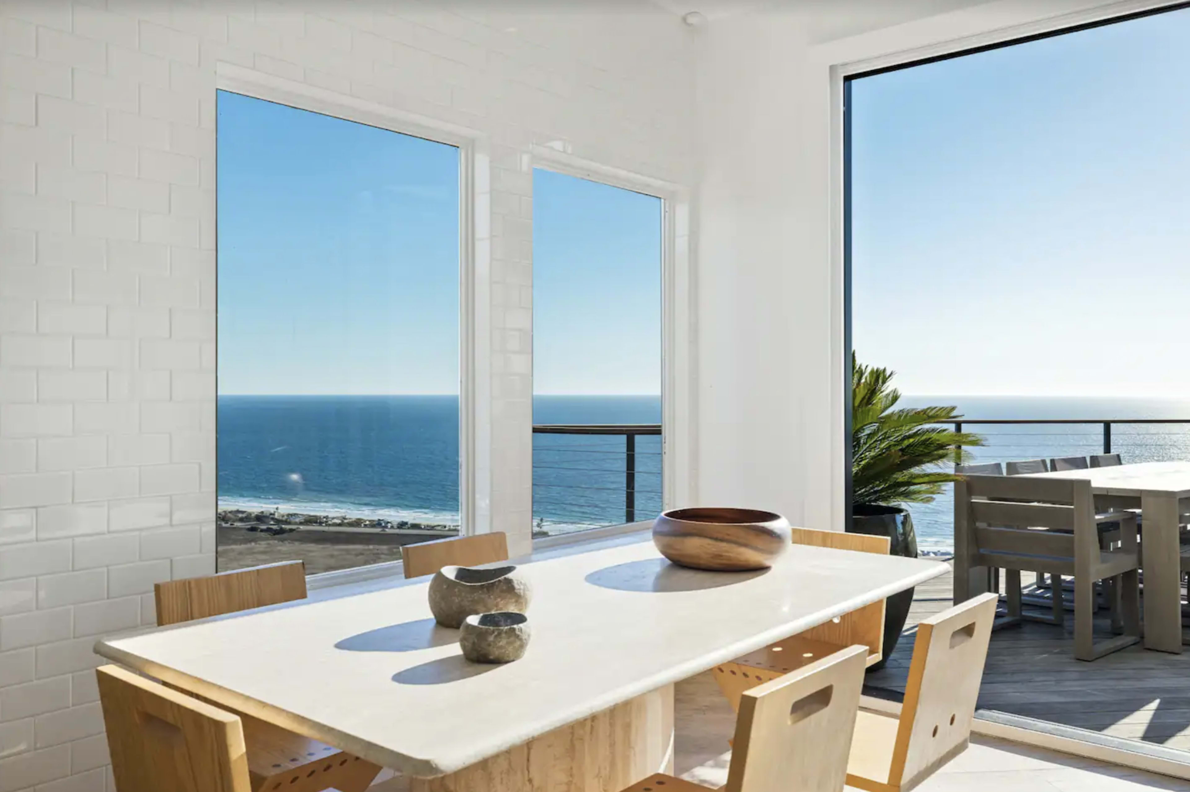 A dining table with wooden chairs is set near large windows overlooking the ocean.