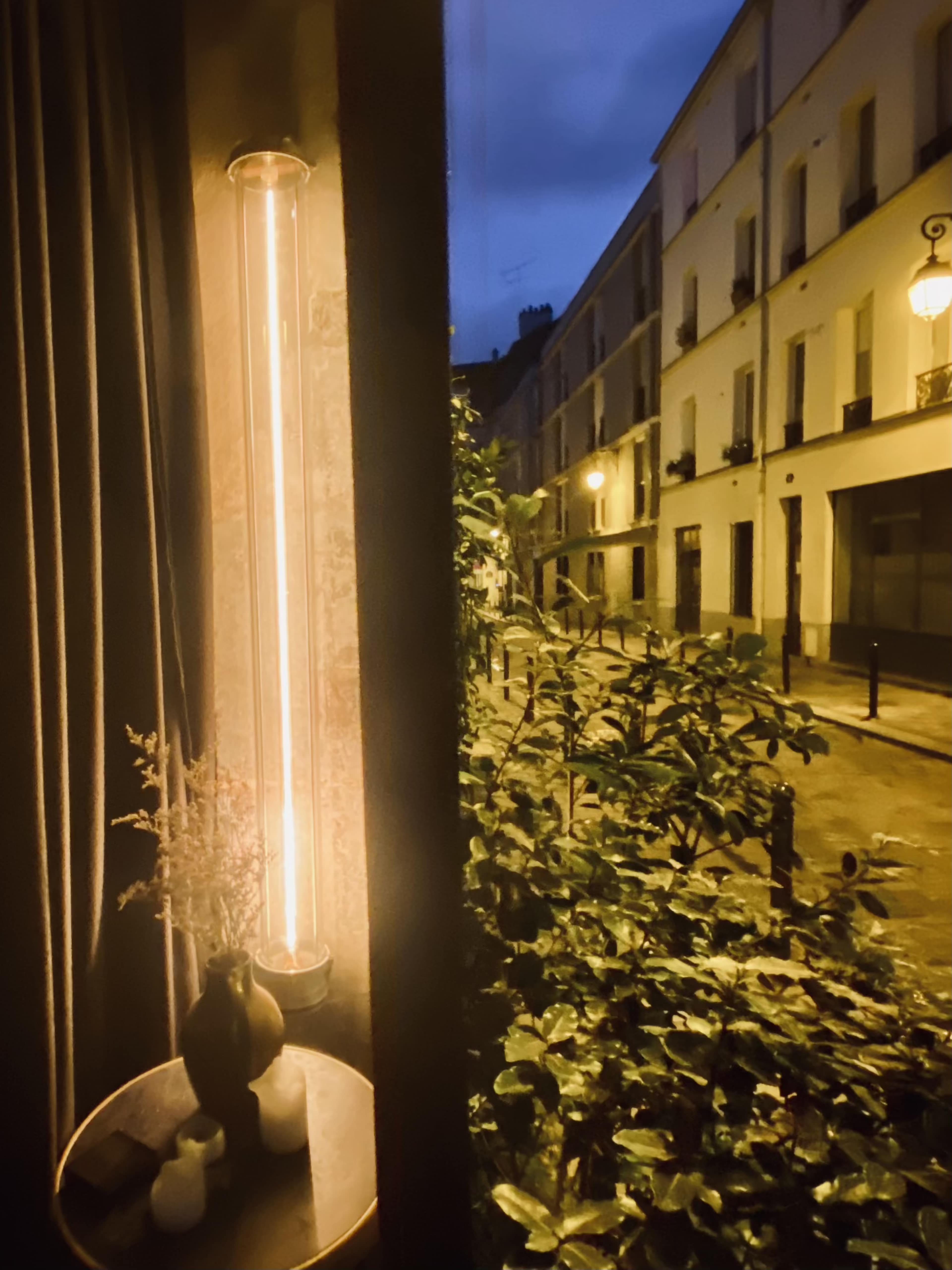 A tall, tubular light fixture illuminates a small table with a vase, while a quiet street lined with buildings is visible outside through the window.