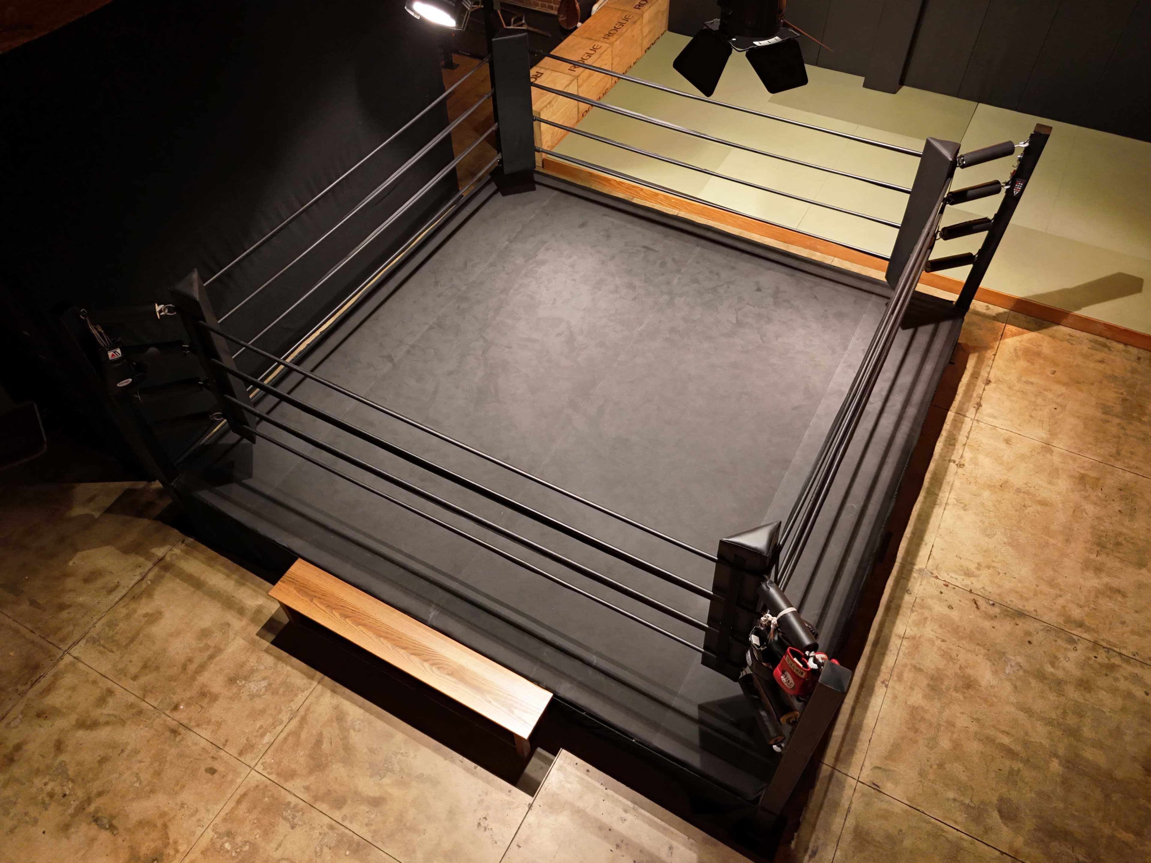 A boxing ring with black padding and ropes is set up in a dimly lit indoor space.